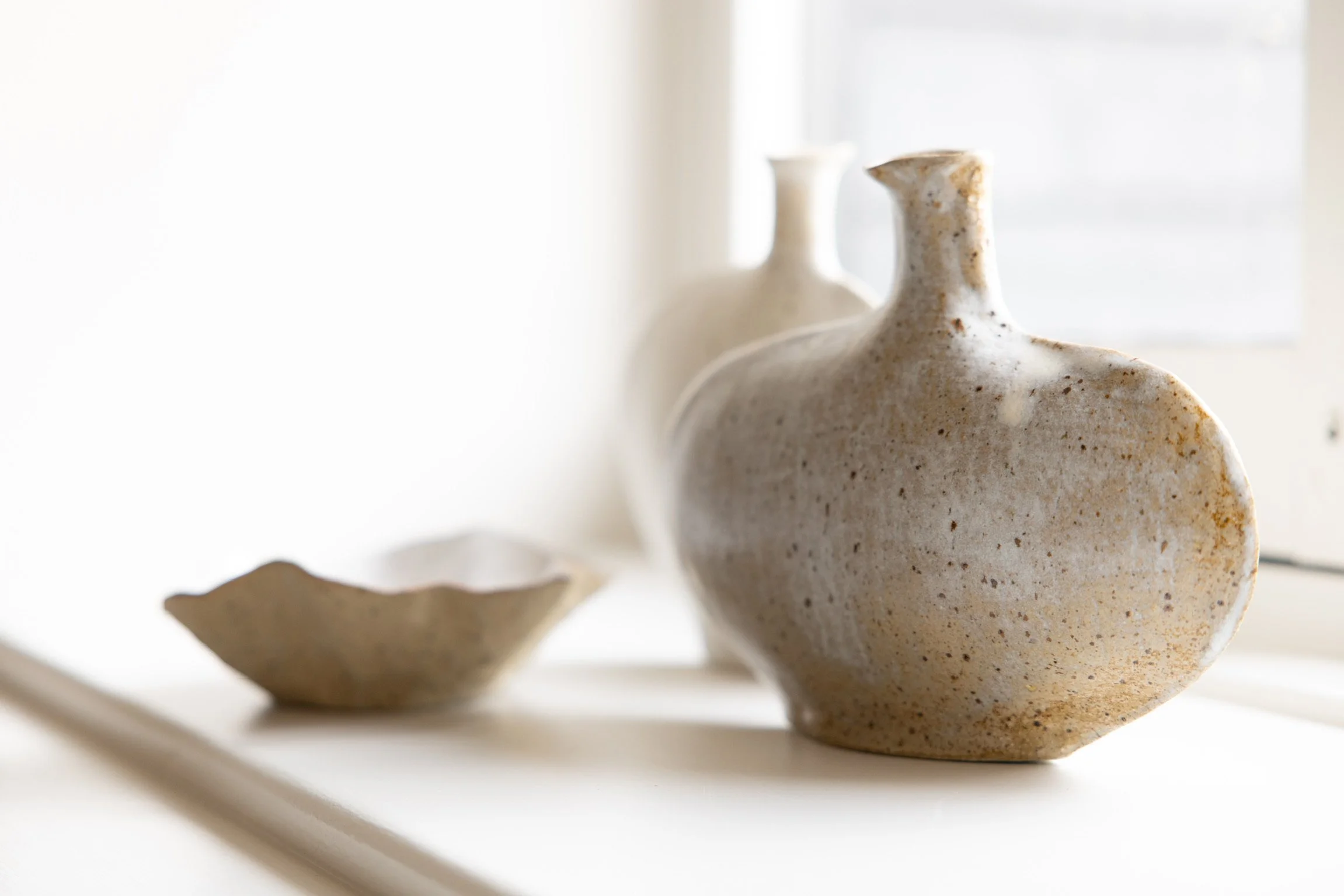 Close-up of a beige ceramic vase with a narrow opening, placed on a white surface with a blurred background and partial clay bowl in the foreground.