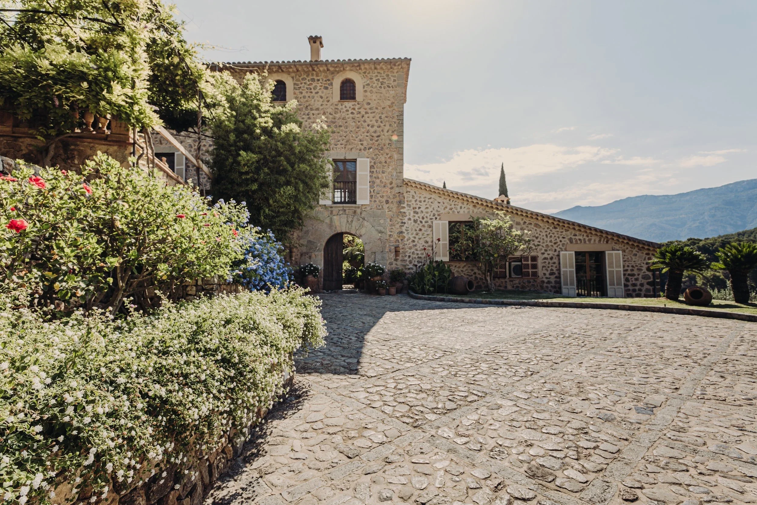 A stone house with white shutters, surrounded by colorful flowers and greenery, with a stone-paved courtyard and a mountain range in the background.