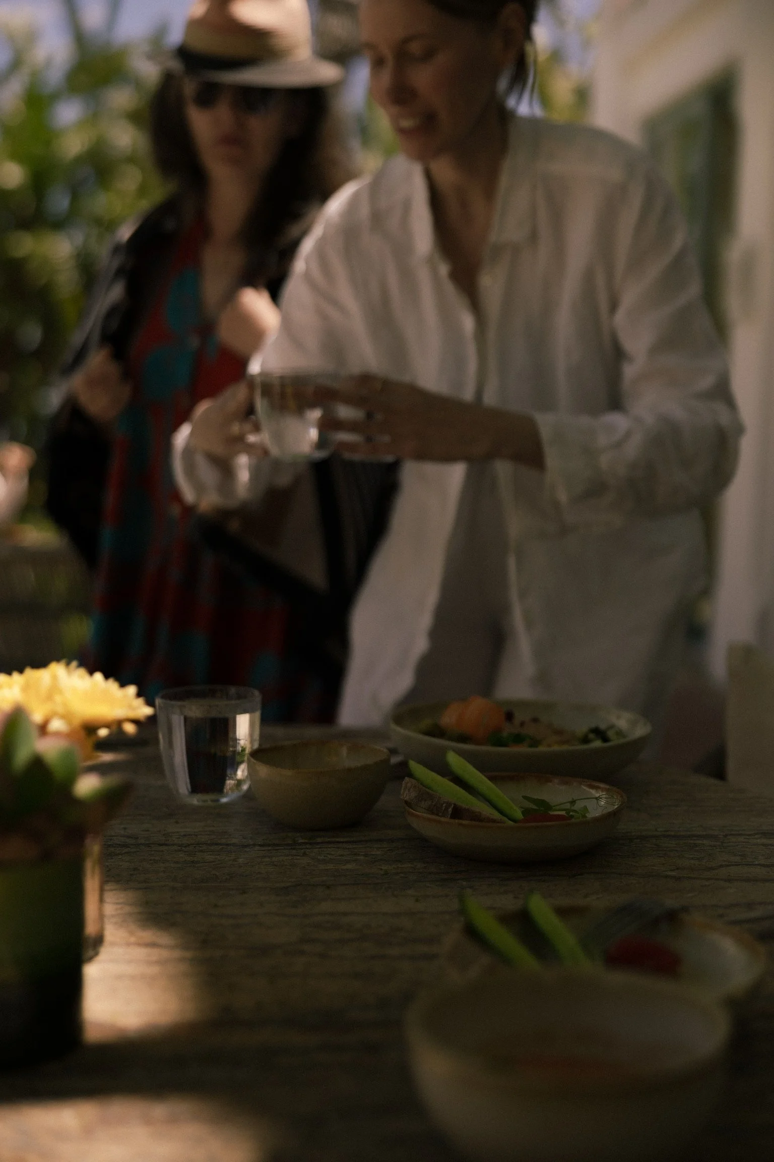 Two women, one with glasses and a hat, and the other in a white shirt, are looking at a tablet at a kitchen or dining table filled with dishes and food. The scene is dimly lit with natural light.