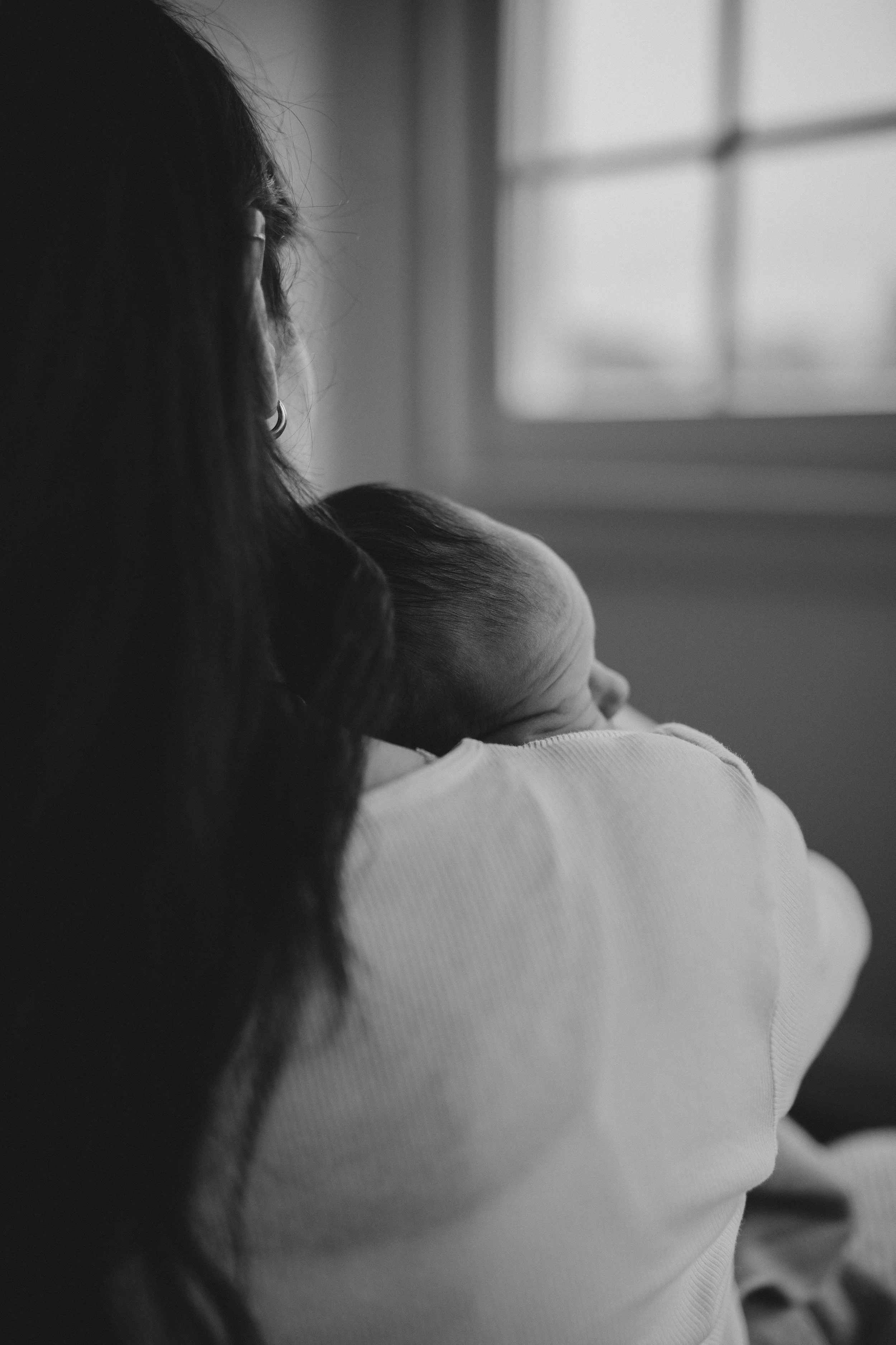 Schwarz-weiß-Fotografie einer Person, die ein Baby auf der Schulter trägt, Blick nach draußen durch ein Fenster.