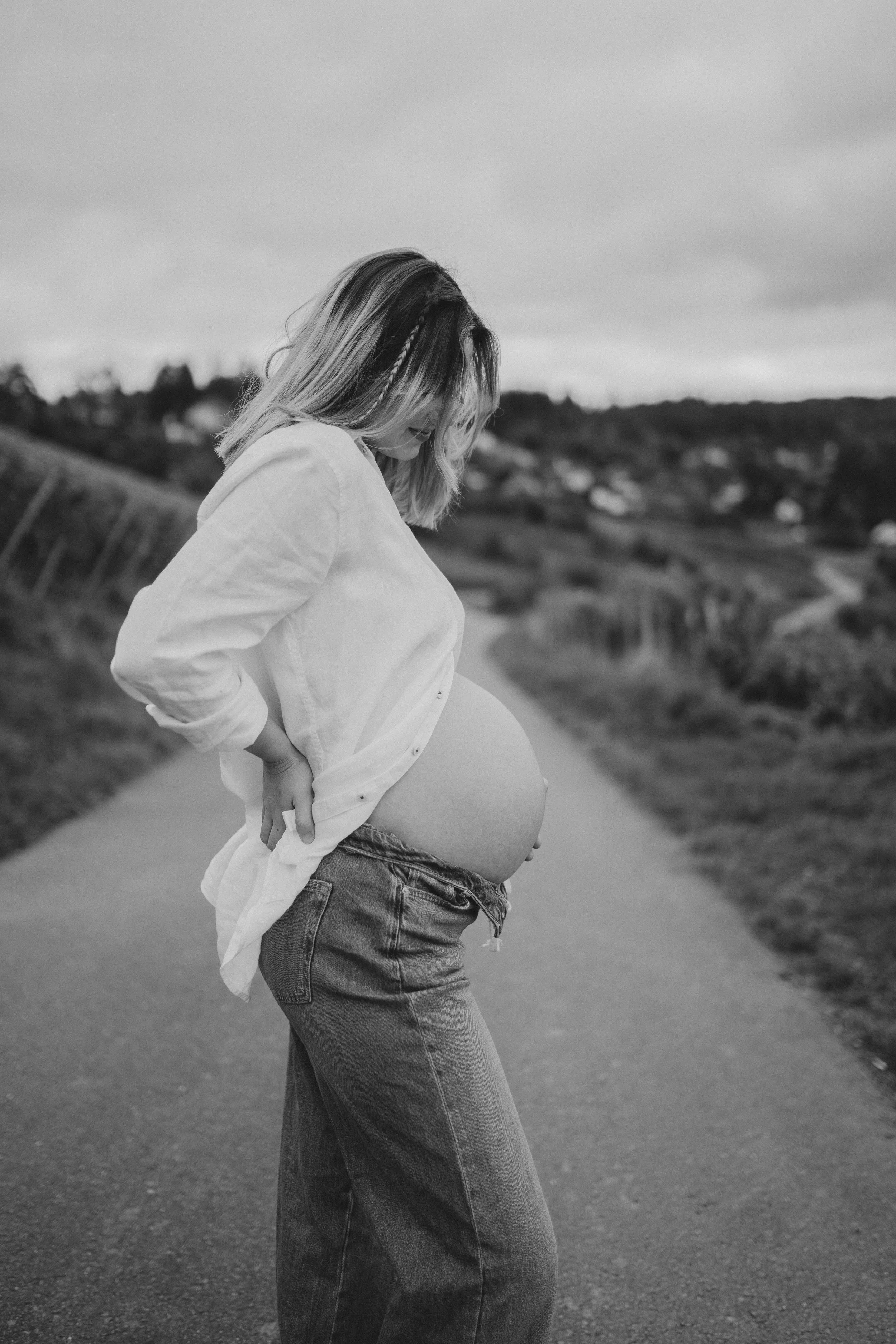Schwangere Frau mit langer Haar, die in Jeans und weißem Hemd steht, Blick nach unten, im Freien auf einer Landstraße.