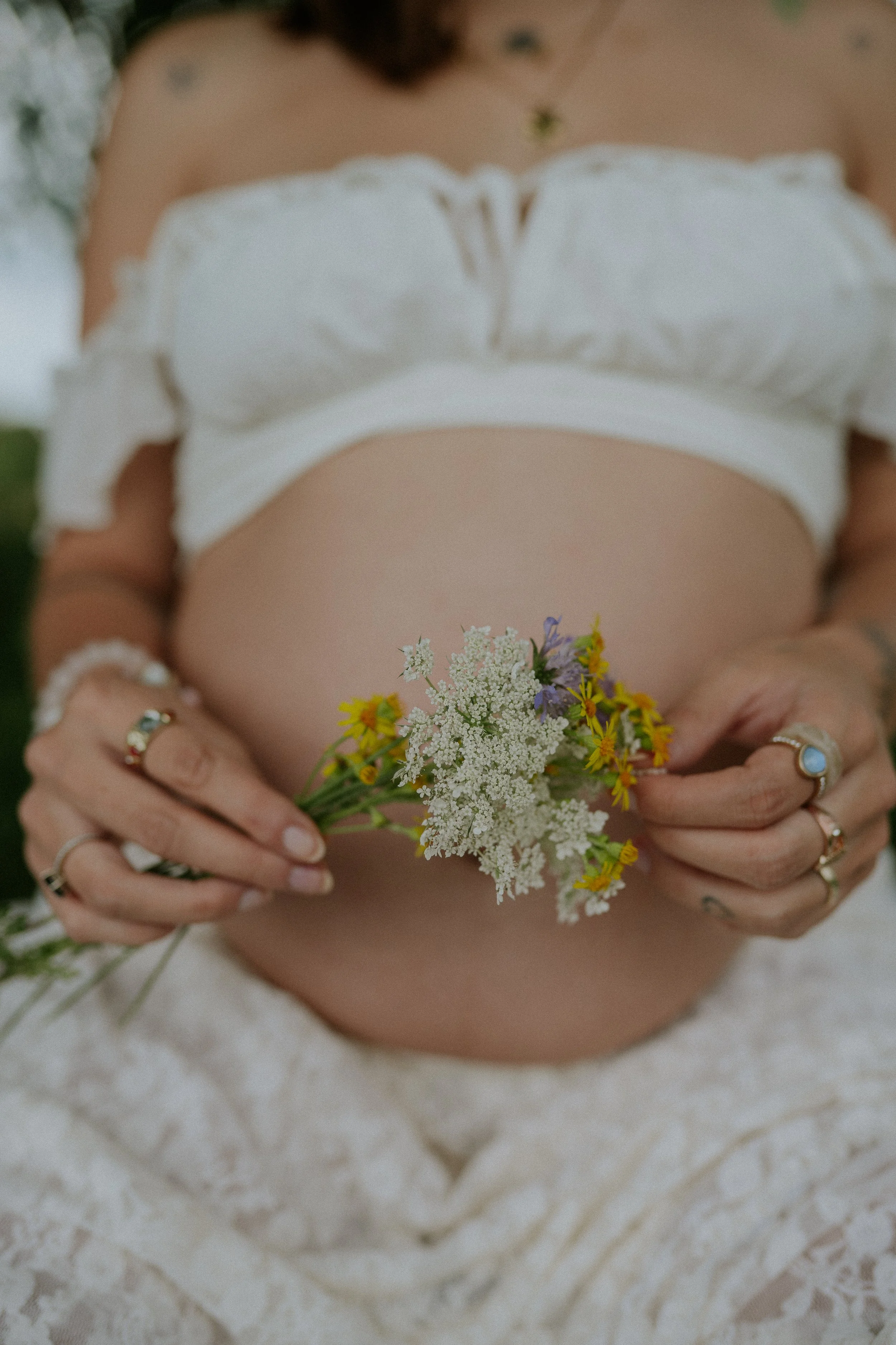 Nahaufnahme einer Frau, die Blumen hält, mit einem weißen Kleid, verschiedene Ringe an den Fingern, im Hintergrund unscharf, weiches Licht.