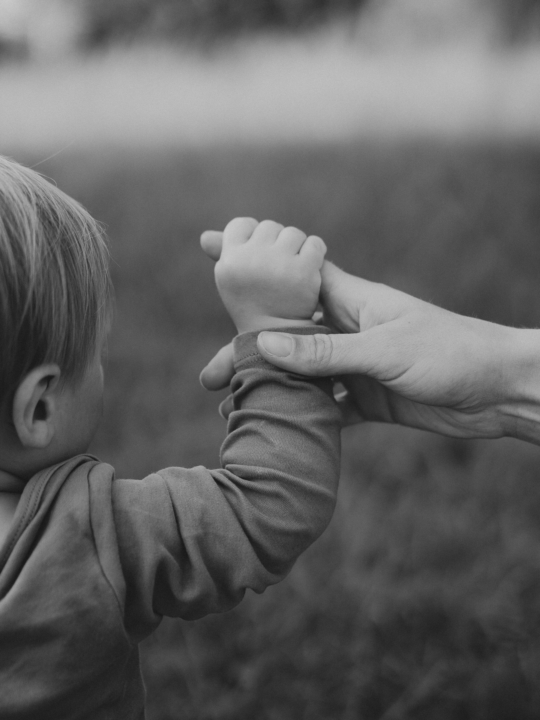 Ein Erwachsener hält die Hand eines kleinen Kindes, beide Hände sind verbunden, im Hintergrund ist unscharf ein Naturfeld. Das Bild ist in Schwarzweiß gehalten.
