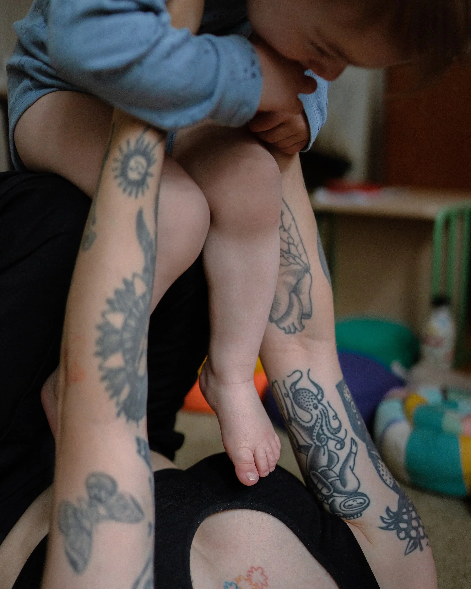 Nahaufnahme von einem Baby, das an einem Erwachsenen geknuddelt wird, mit sichtbaren Tattoos auf den Armen des Erwachsenen.
