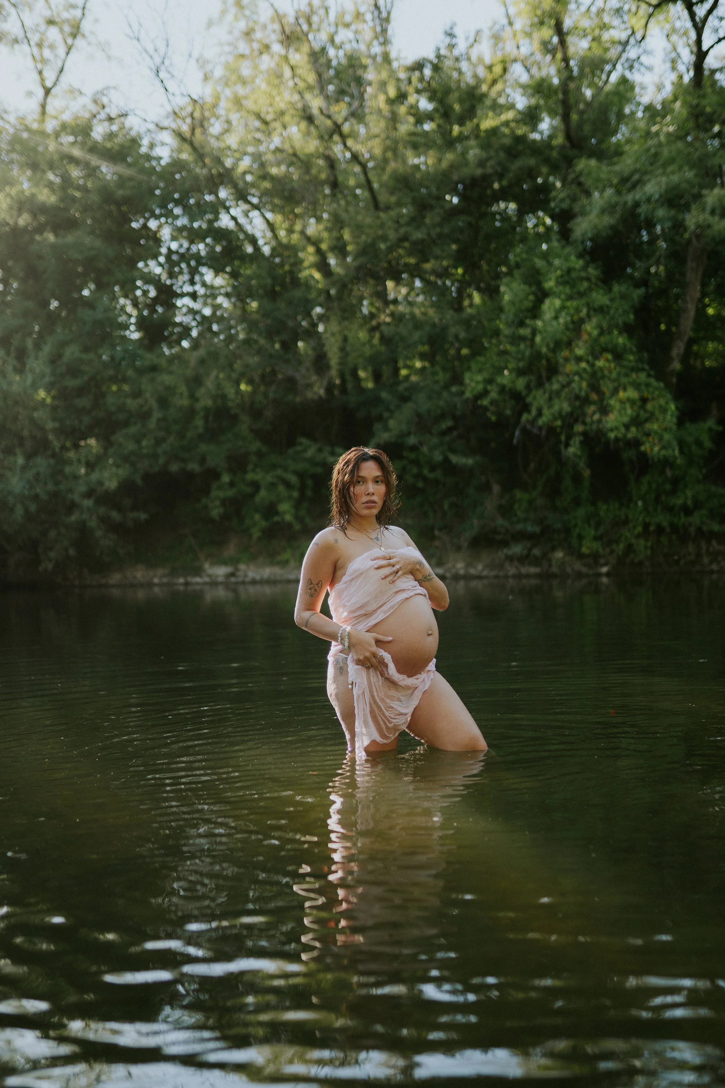 Eine schwangere Frau steht im Wasser eines Flusses oder Sees, umgeben von grünen Bäumen, bei Sonnenuntergang, trägt ein leichtes, enganliegendes Kleid. Sie blickt nach vorne, ihre Hände liegen auf ihrem Bauch, der Wasserstand reicht bis zu ihren Ober
