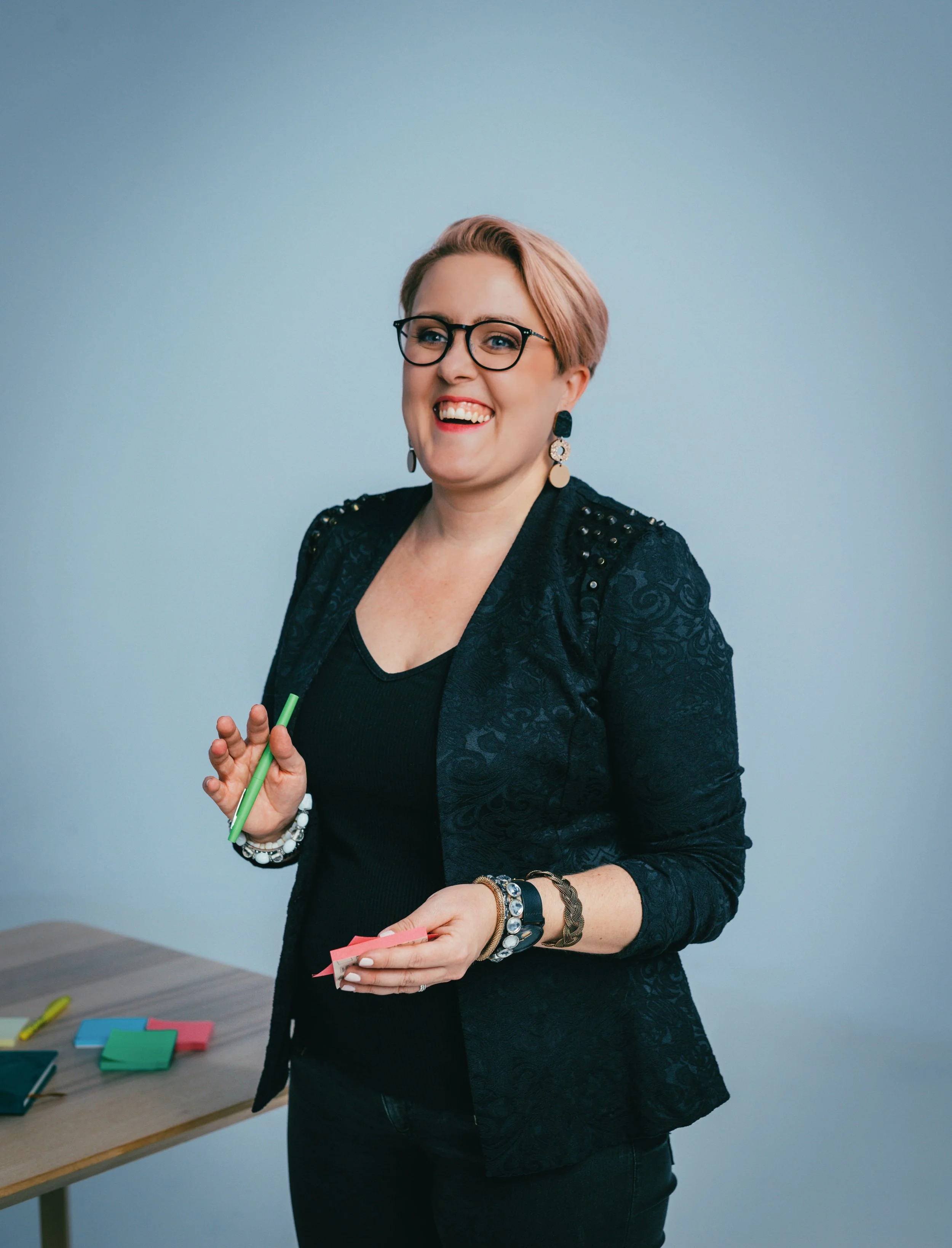 A woman with short, light brown hair, glasses, and earrings, smiling and holding colorful sticky notes and a pen, standing next to a table with more sticky notes on it.