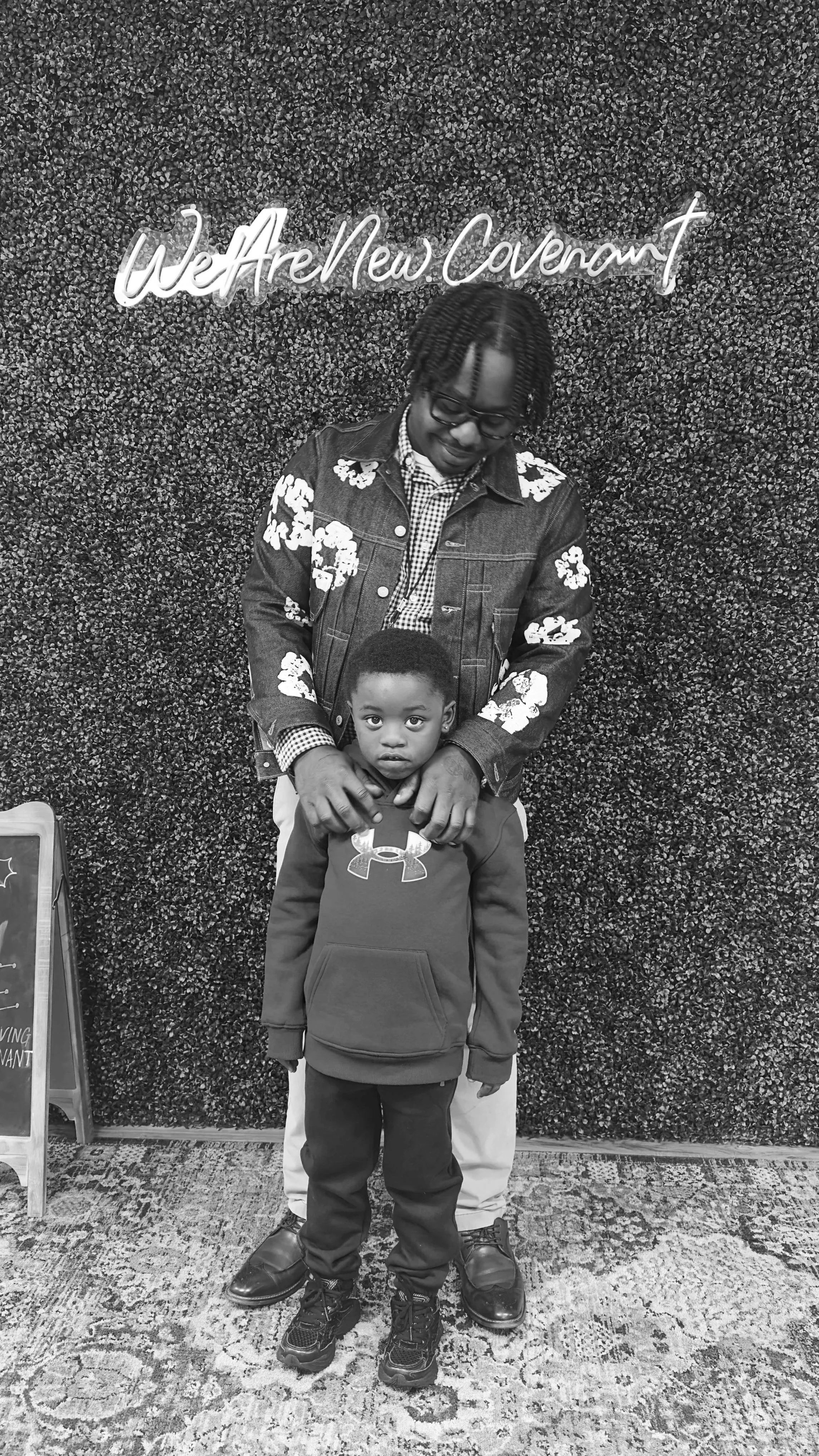 A man with glasses and braided hair holding a young boy in front of a textured wall with a neon sign that reads 'WeAreNewCovenant'. The boy is wearing a hoodie with an Under Armour logo, and the man is wearing a floral jacket and checkered shirt.