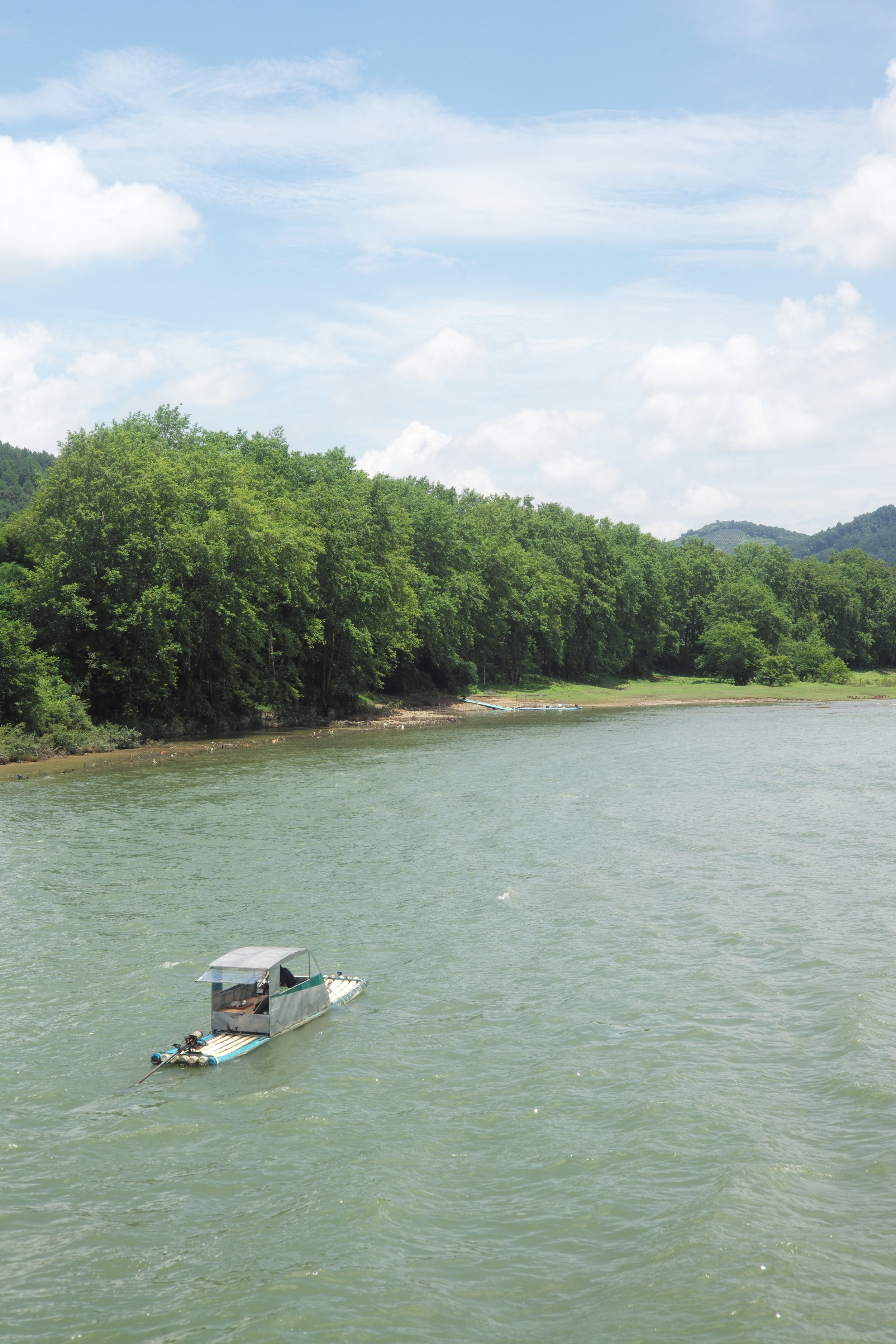 A small boat with a canopy floating on a river with green hills and trees in the background under a partly cloudy sky.
