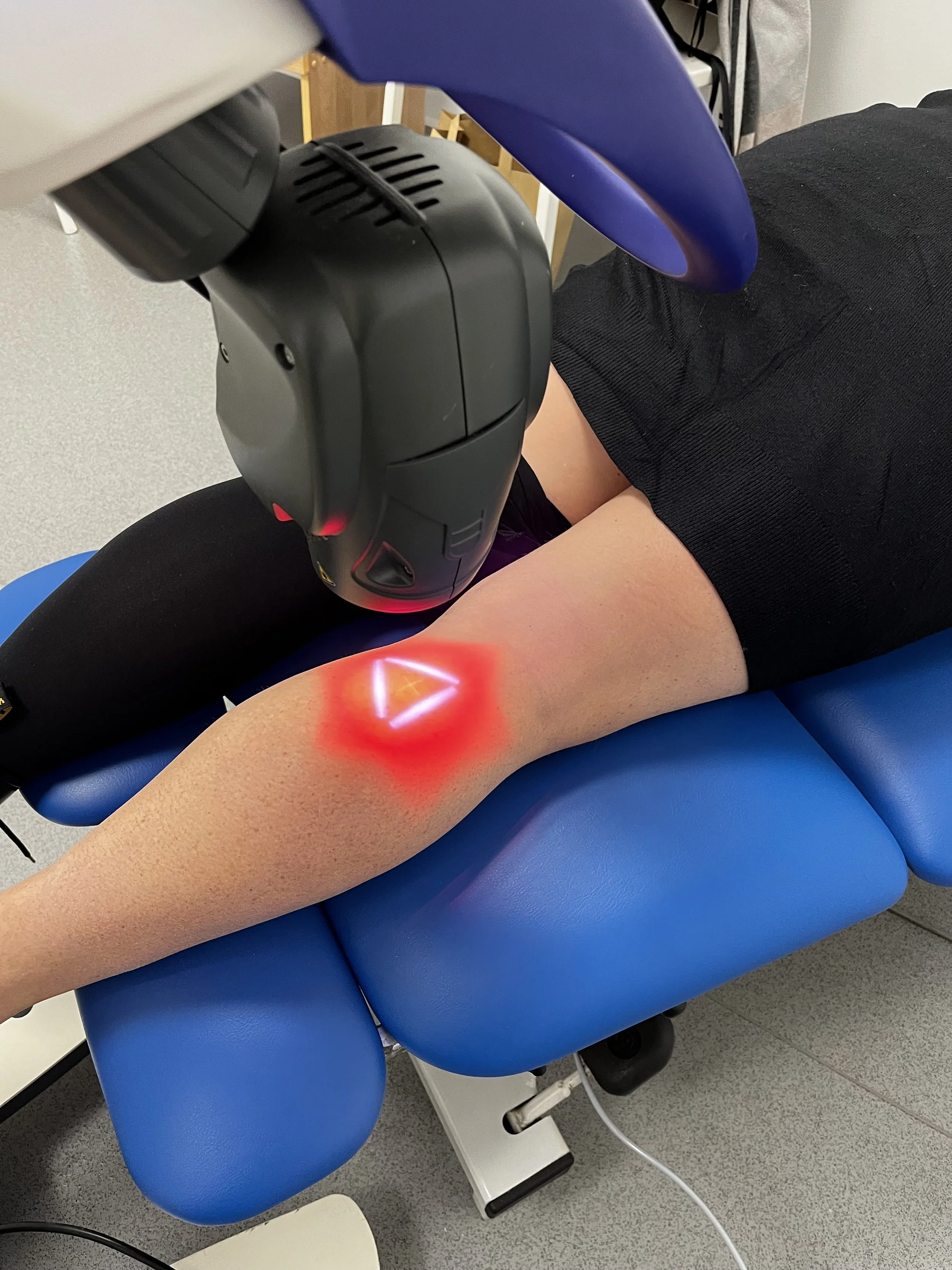 A laser device emitting red light onto a person's leg, who is lying on a blue medical chair in a clinical setting.