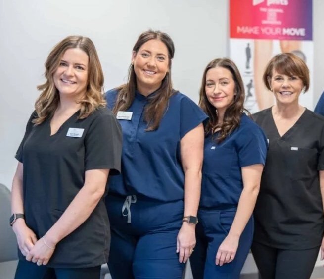 Four smiling healthcare workers in scrubs standing side by side in a hospital or clinic setting.