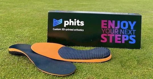 Pair of orange and black custom 3D-printed orthotic shoe insoles on grass with packaging box in the background that reads 'phits' and 'Enjoy your next steps'