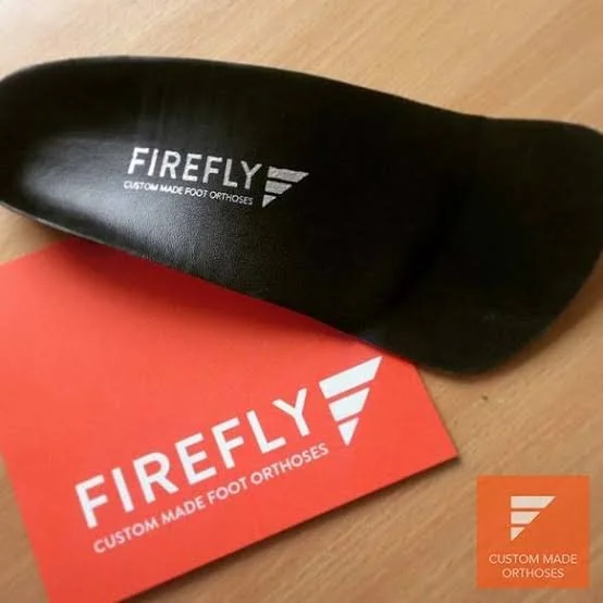 A black shoe insole with the words 'FIREFLY CUSTOM MADE FOOT ORTHODES' printed on it, resting on a red card that has the same branding and a logo with three horizontal bars.