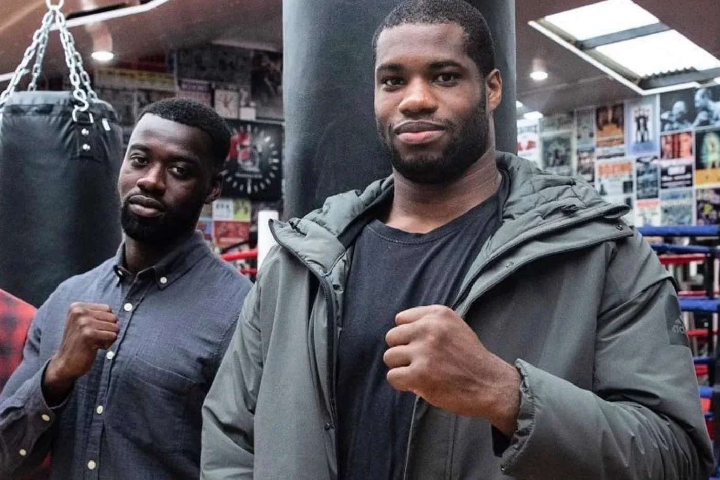 Two men standing inside a boxing gym, both with fists raised in a fighting stance. The man on the left is wearing a gray button-up shirt, and the man on the right is wearing a black t-shirt and a gray jacket. The background shows a punching bag and various boxing-related posters.