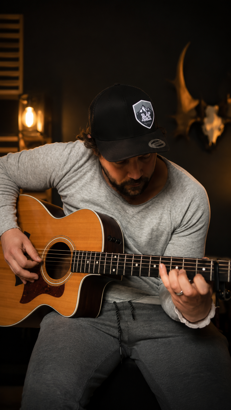 Man som spelar akustisk gitarr i ett mörkt rum med en väggdekoration av ett djurhuvud.