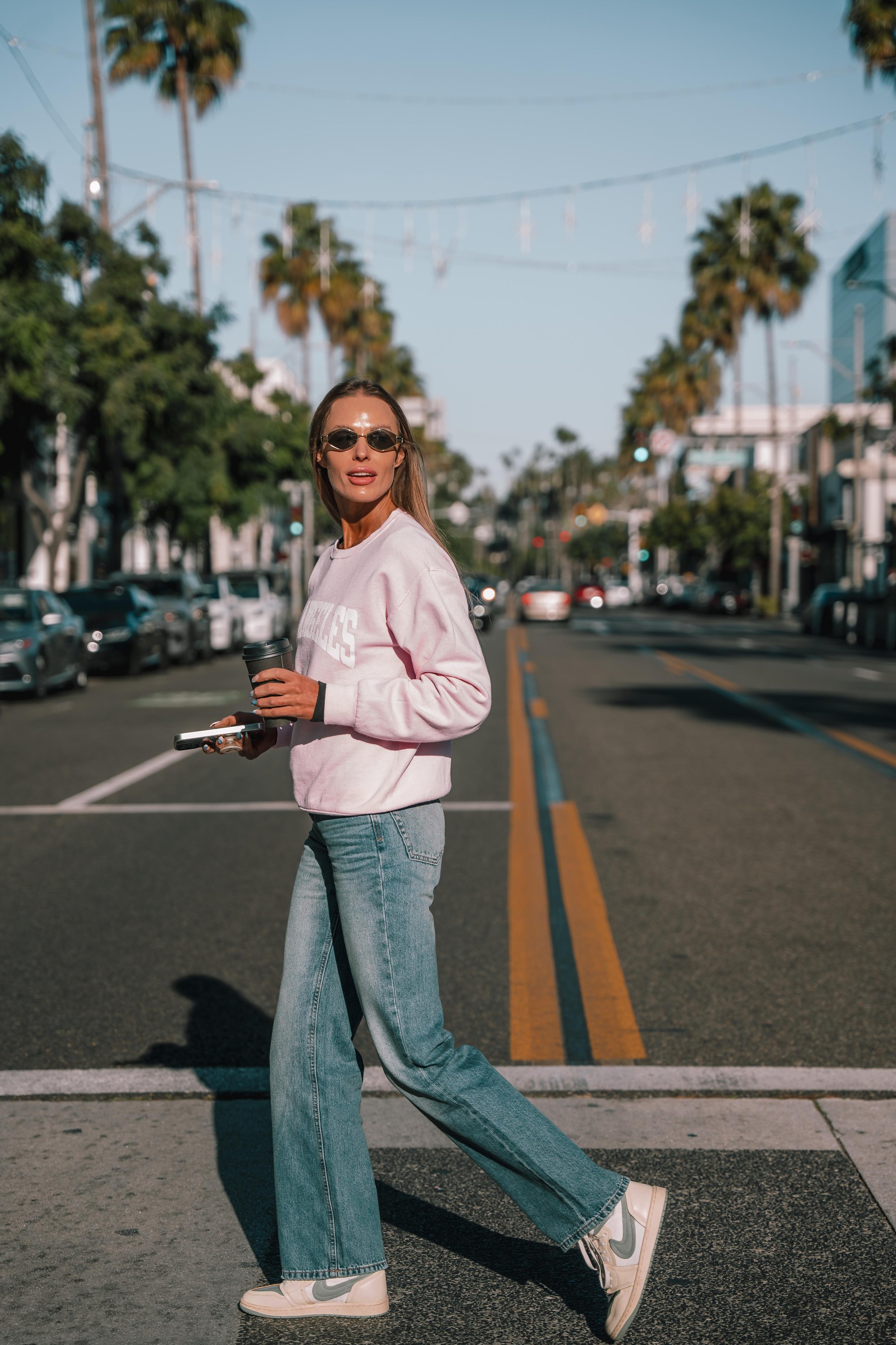 Woman crossing a city street holding a coffee cup and a phone, wearing sunglasses, a pink sweatshirt, blue jeans, and sneakers, with palm trees and storefronts in the background.