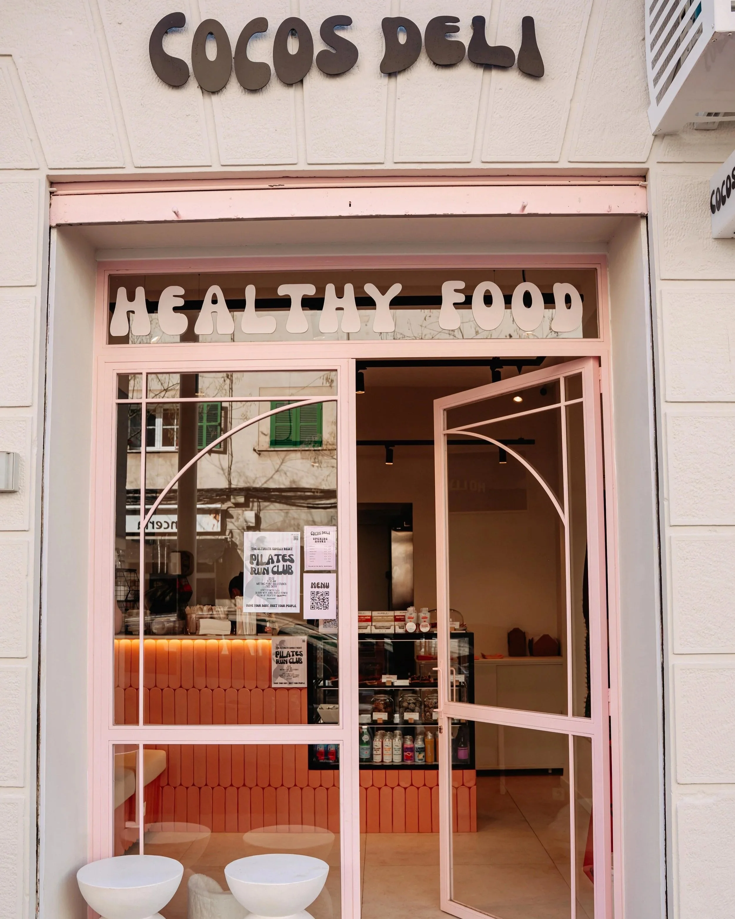 Front view of a health food and smoothie shop named Coco's Deli with a pink storefront and large glass windows. Inside, there are signs advertising Pilates and a run club, and various health products on display. The shop features a clean, modern design with white walls and seating outside.