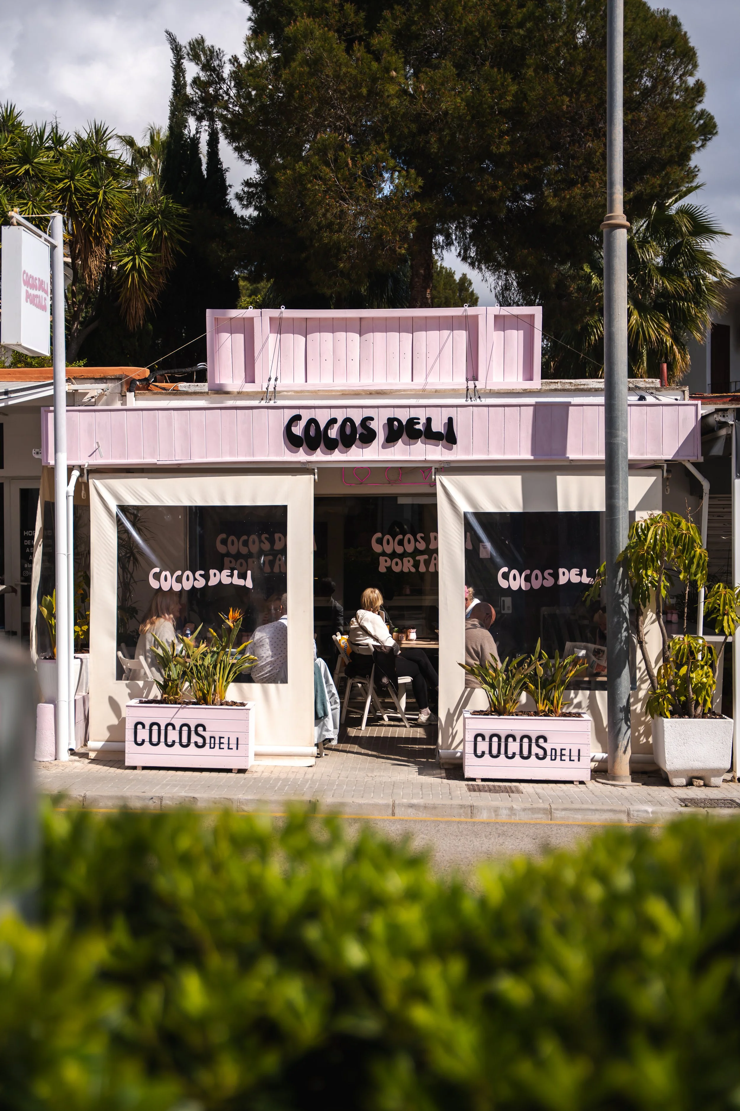 Pink storefront of Cocos Deli with outdoor seating area and potted plants, sign with black lettering, trees and cloudy sky in background.
