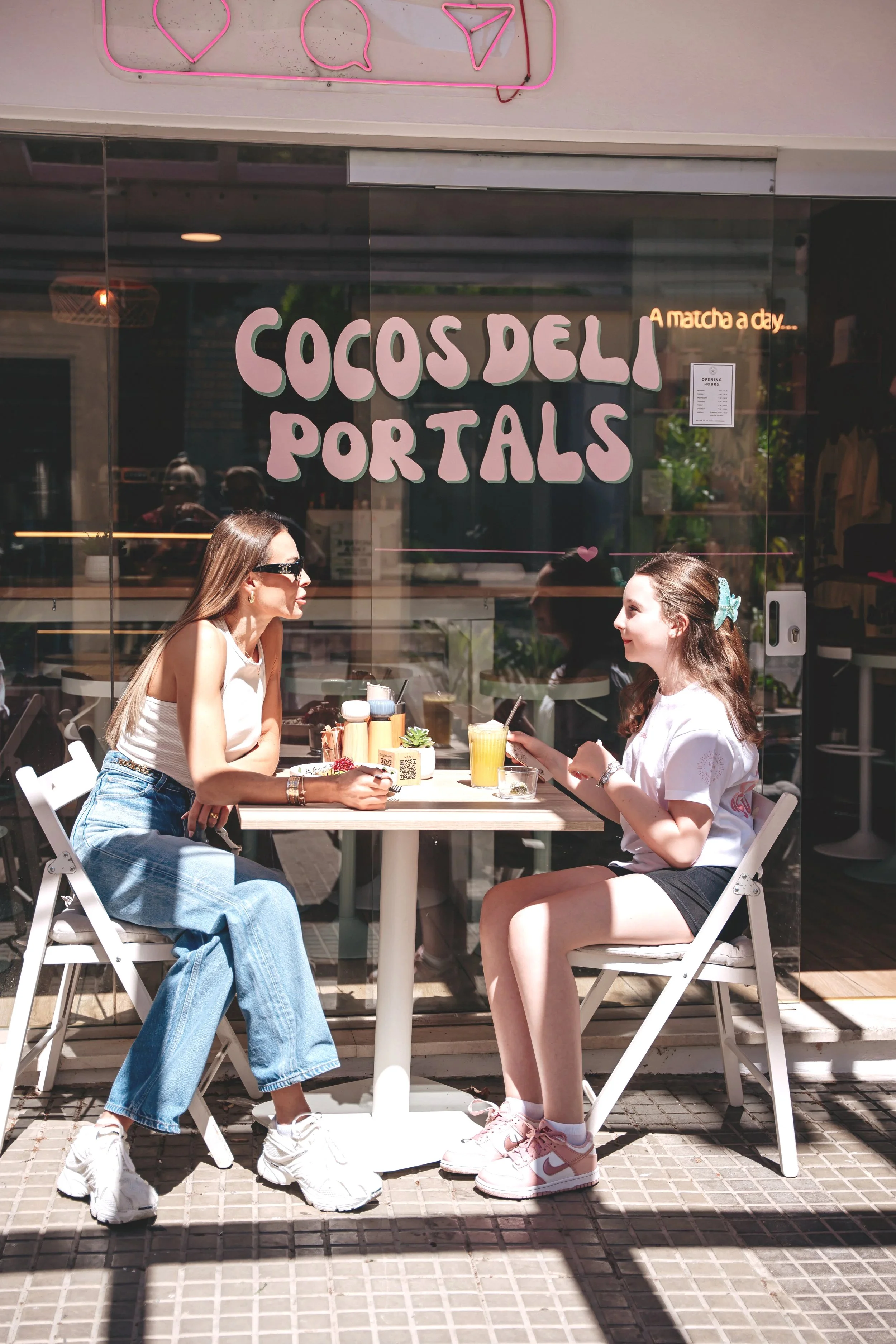 Two women sitting at an outdoor table outside a cafe called "Cocos Deli Portals" on a sunny day. They are having drinks and chatting. The cafe has pink neon signs above the window.