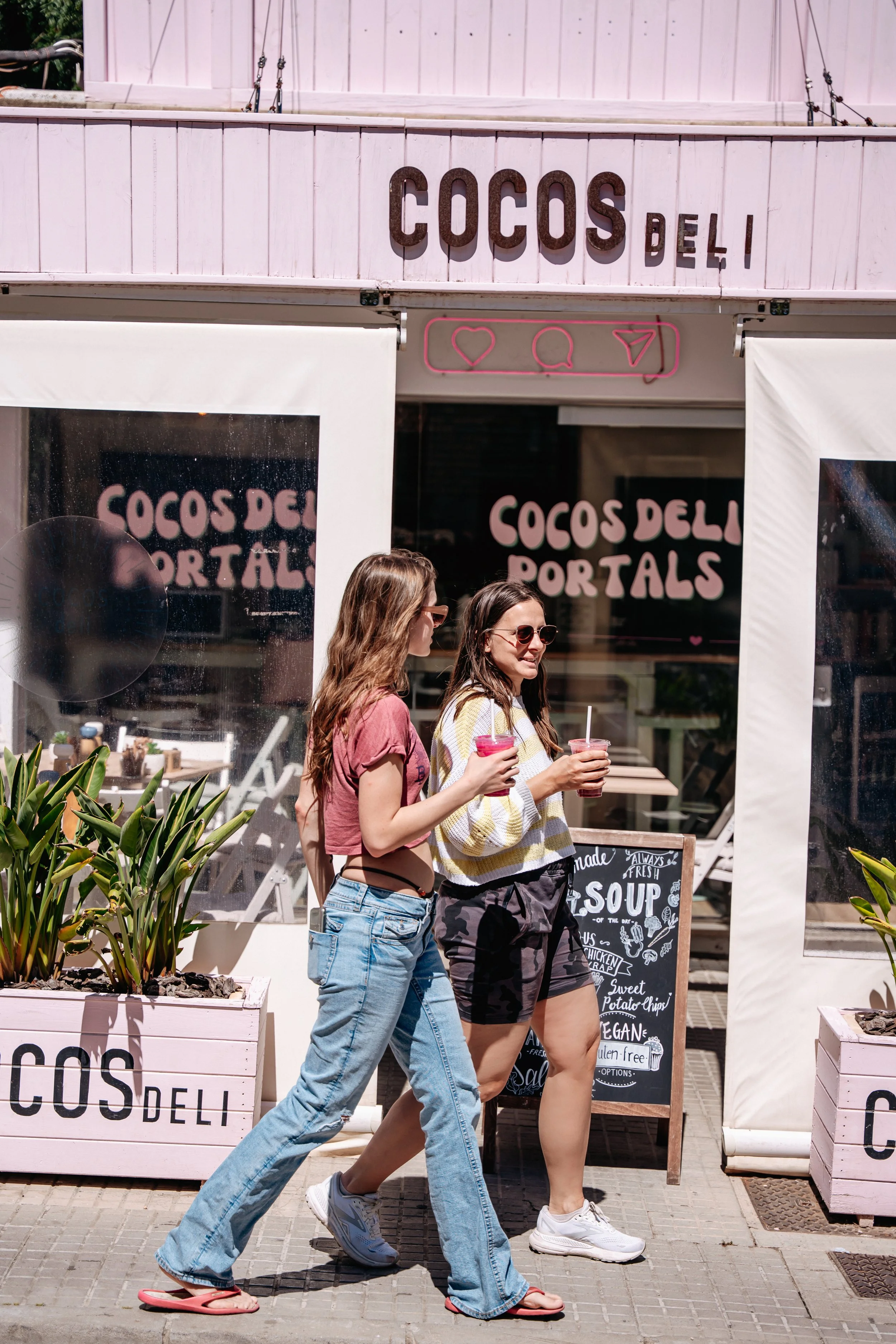 Two young women walking past a pink storefront with signs that say 'Cocos Deli' and 'Cocos Deli Portals,' holding pink drinks with straws.