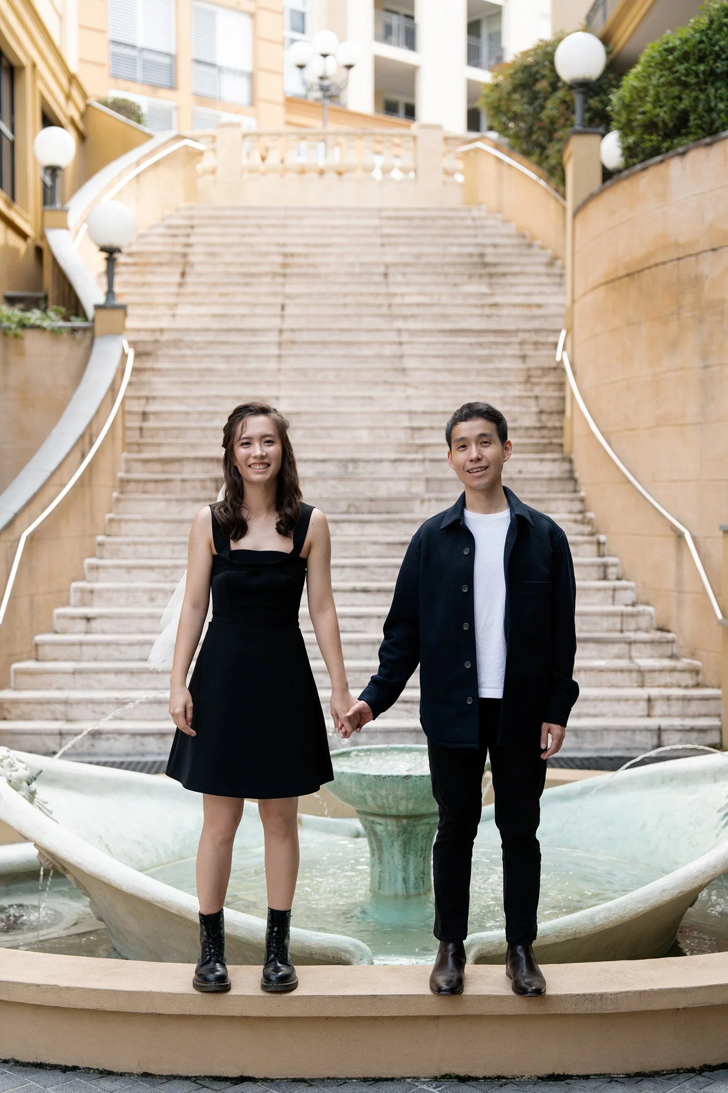 Paul and Krystel holding hands stand in front of the fountain look at the Brisbane wedding photographer in Emerald Lakes Village