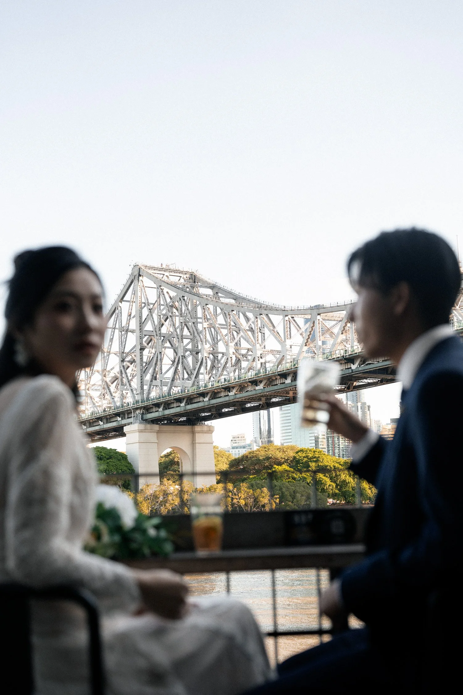 Brisbane story bridge at background, bride and groom sitting next to each other holding a drink in Wilson Outlook Reserve