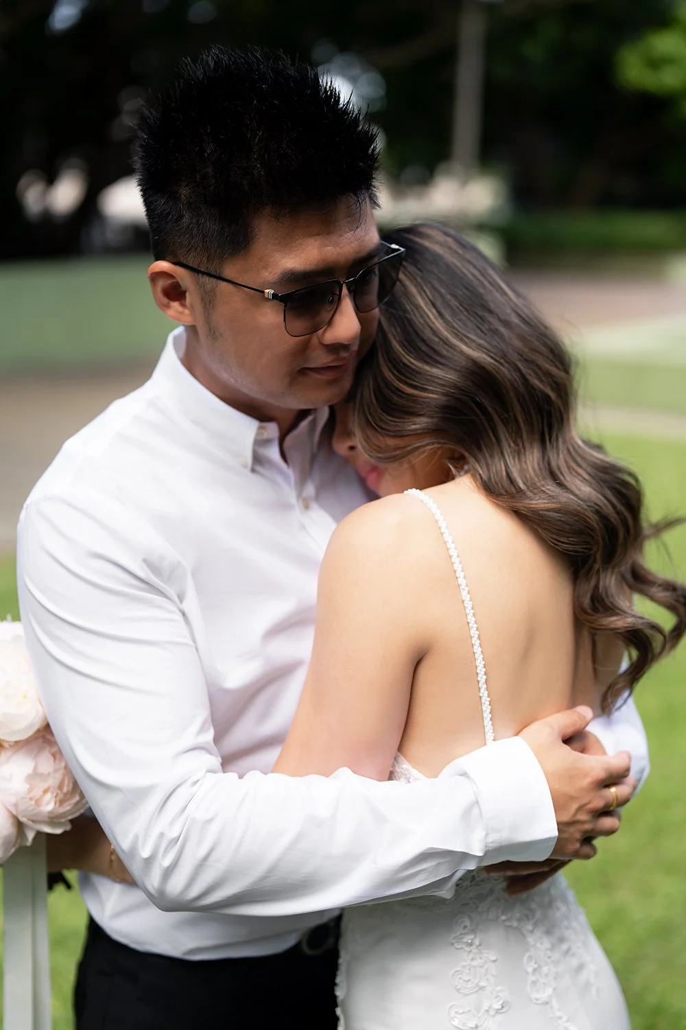 Groom Kai hugging the bride Tiffy as she rests her head gently on his shoulder, lying on the grass in a serene outdoor setting in Queensland. The scene captures a modern artistic touch, with the couple’s relaxed and intimate embrace surrounded by nat