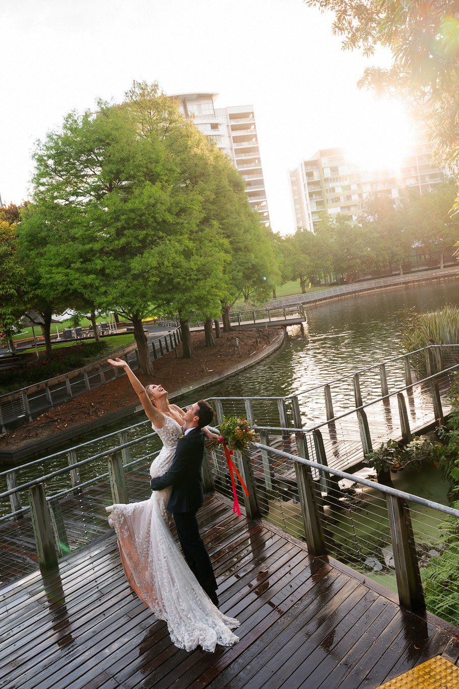bride Sonya raising her hand when groom Giordan lifting her up in Roma street parkland with sun out after running, captured by Brisbane wedding photographer Whistler Li