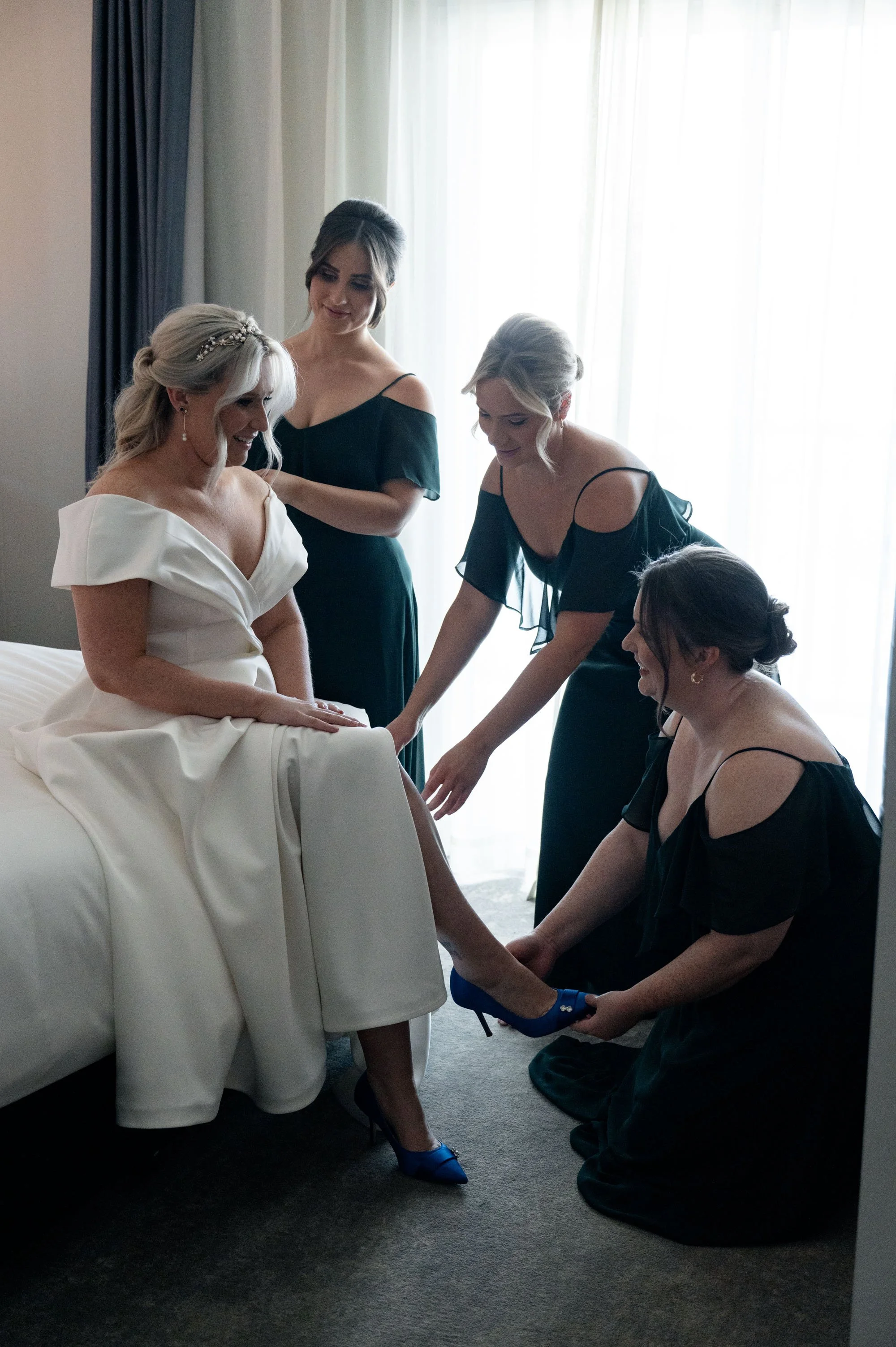 One bridesmaid gently touching the bride's dress while another places a hand on her shoulder, offering support and affection during a special moment before the wedding