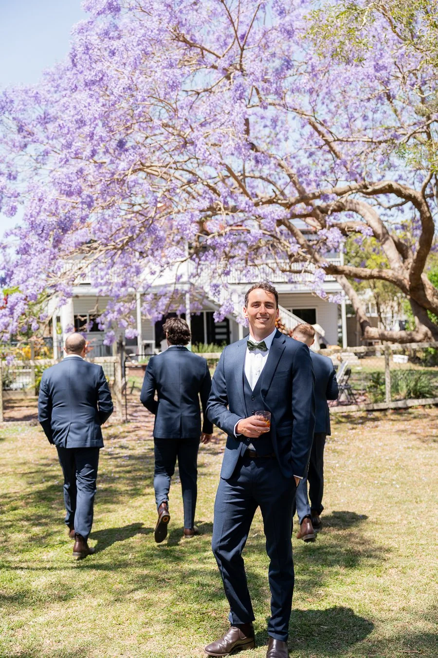 A groomsman walked toward the jacaranda trees with his back to the camera, while the groom turned to face the camera