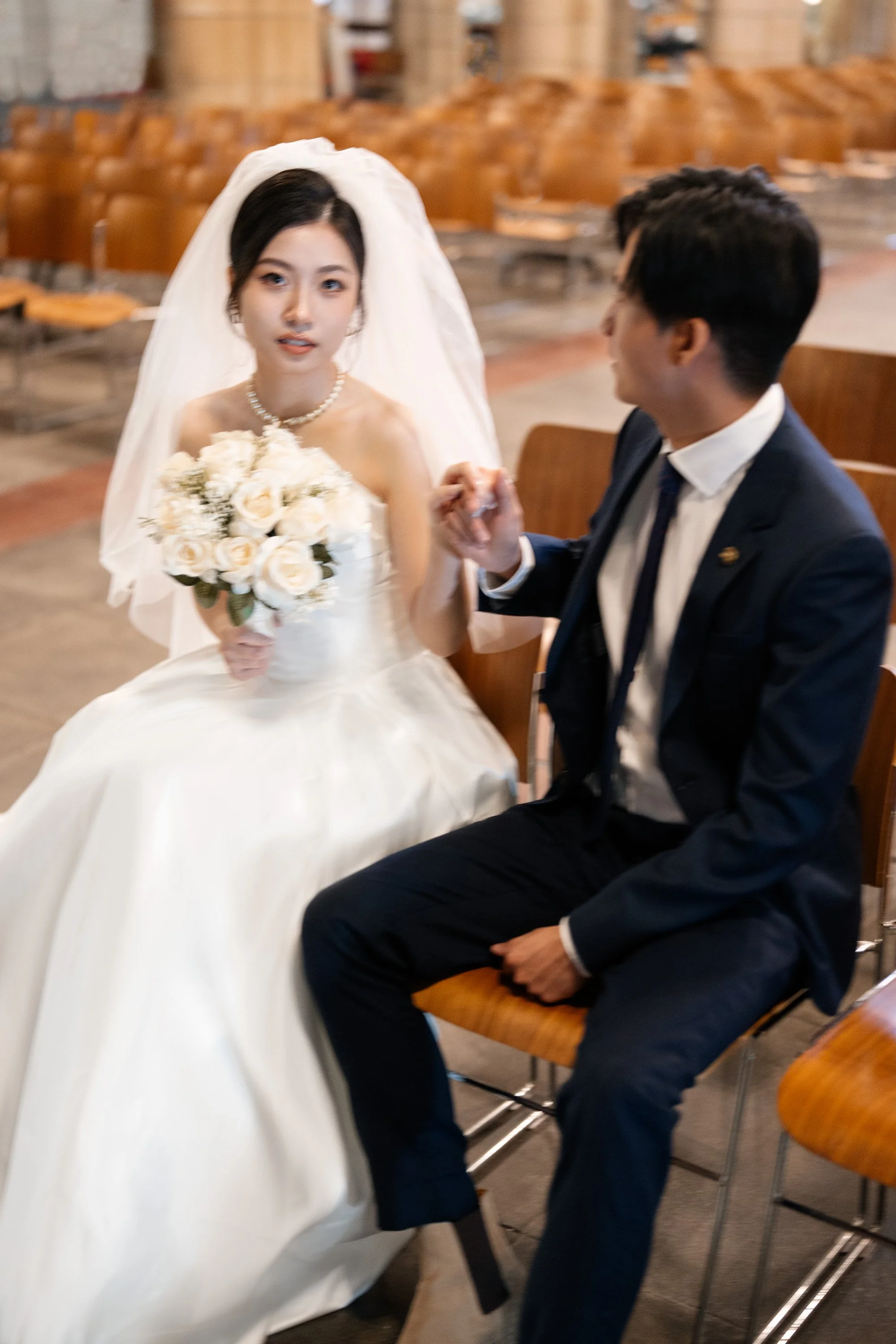 groom holding bride's hand when she look at the camera and they both sitting on the chair