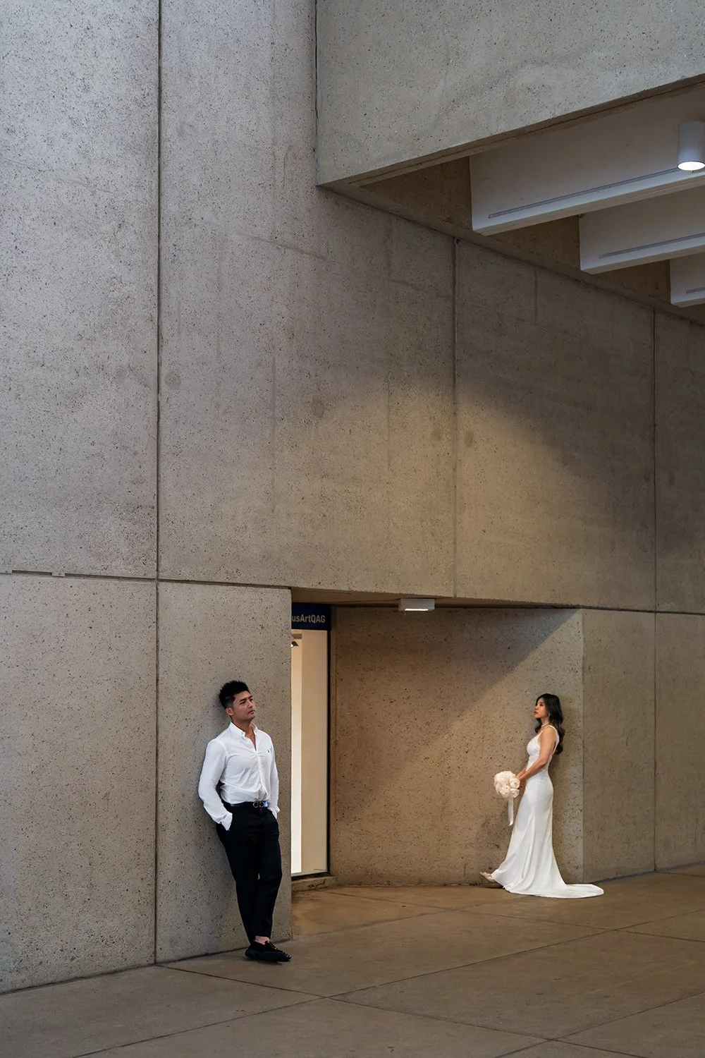 Bride Tiffy leaning gracefully against a wall under warm, soft lighting, while the groom Kai stands beside her, also leaning casually with his head tilted upward in a relaxed pose. Both exude a calm, intimate vibe, with the bride in her elegant weddi