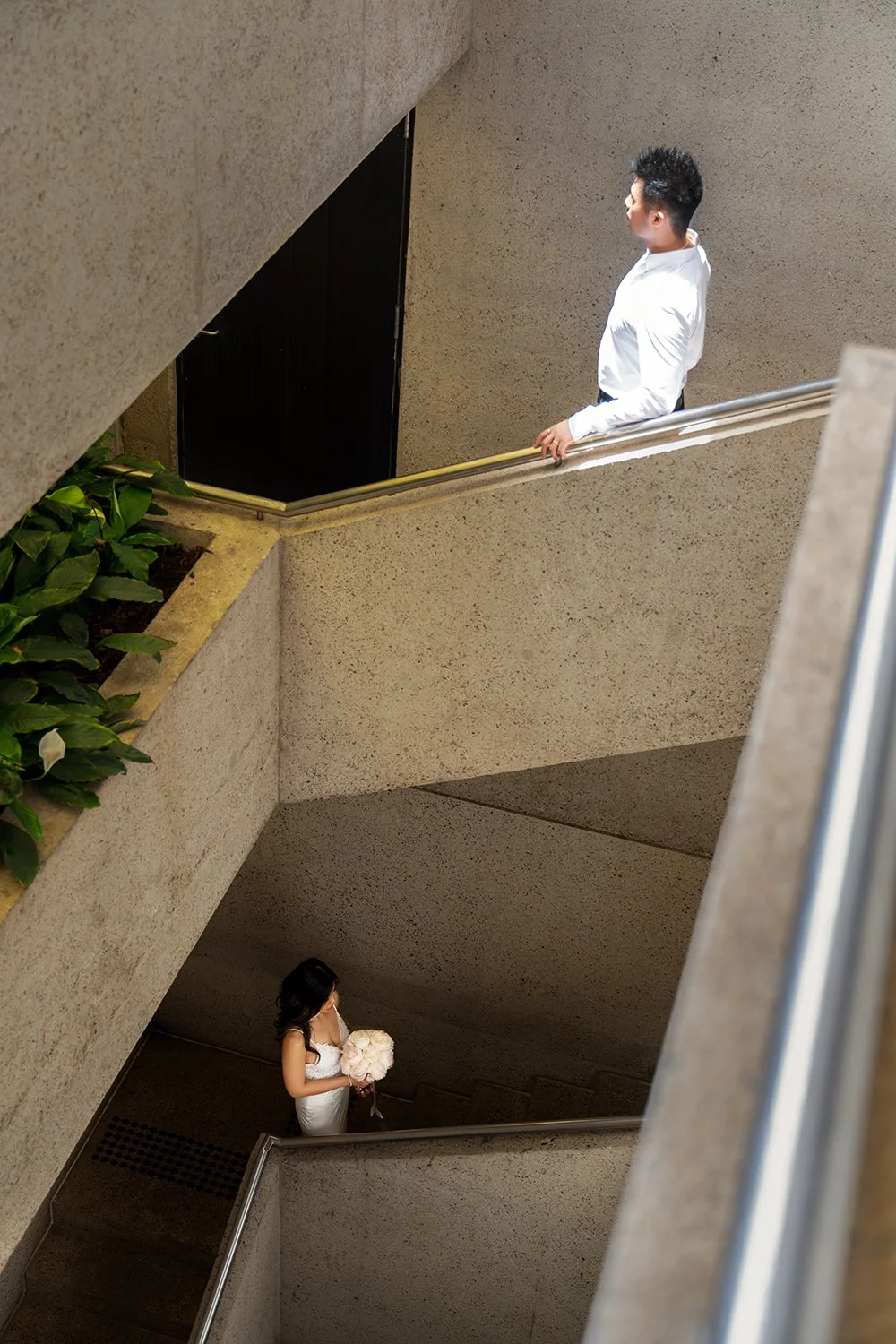 Bride Tiffy and groom kai positioned on intersecting staircases, with the groom standing on the upper level staircase and the bride on the lower level staircase. The architectural lines create a striking composition, highlighting their connection des