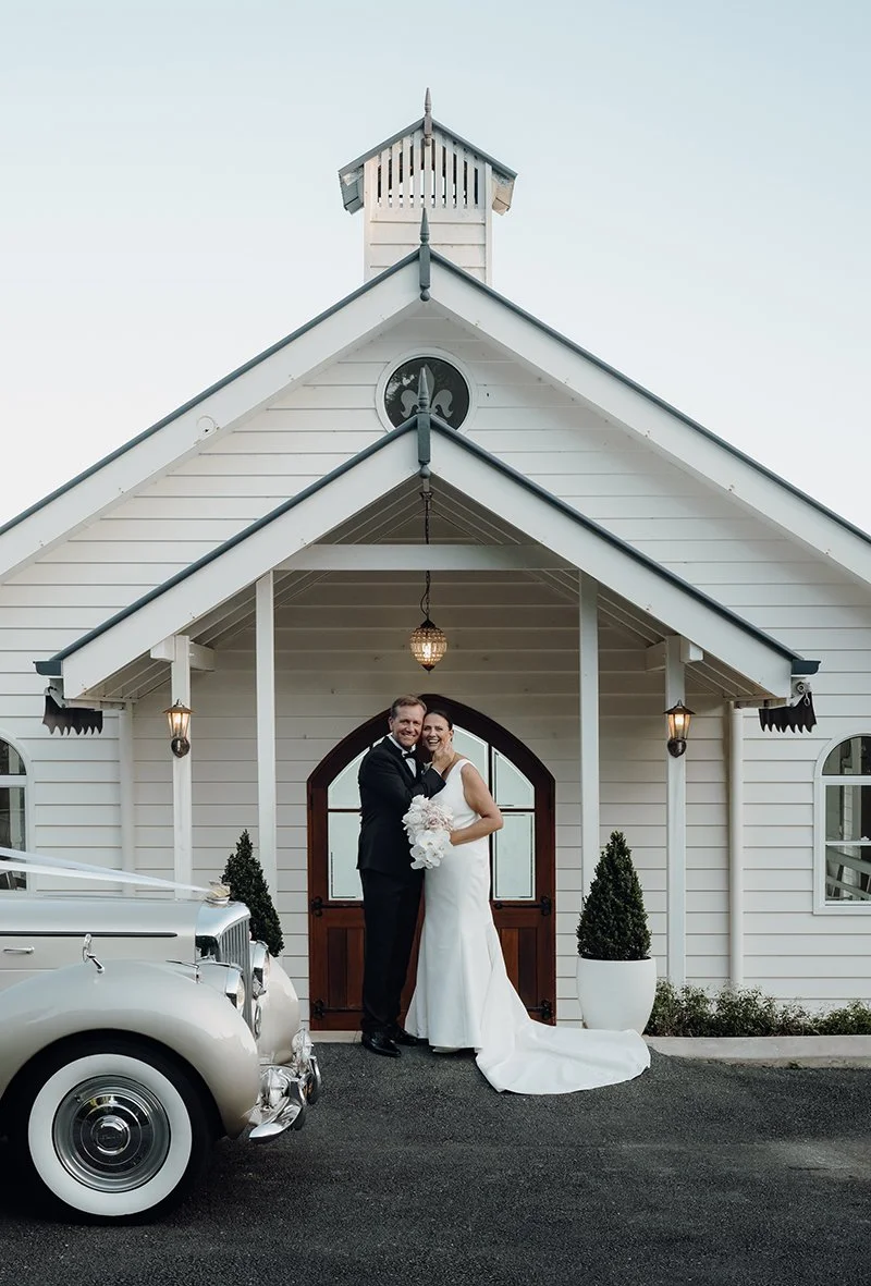 Michelle and Shaun holding each other look at Whistler Li, a old wedding car parking in front of them, and white chapel behind them in Tiffany's Maleny