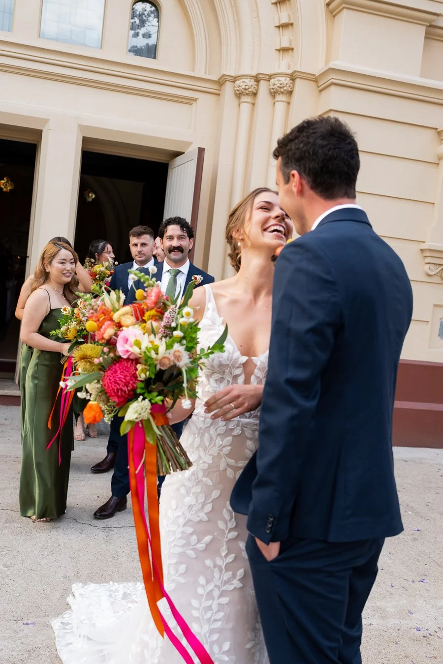 bride Sonya smell to groom Giordan with love and bridal party behind them smell too outside St Mary's Catholic Church