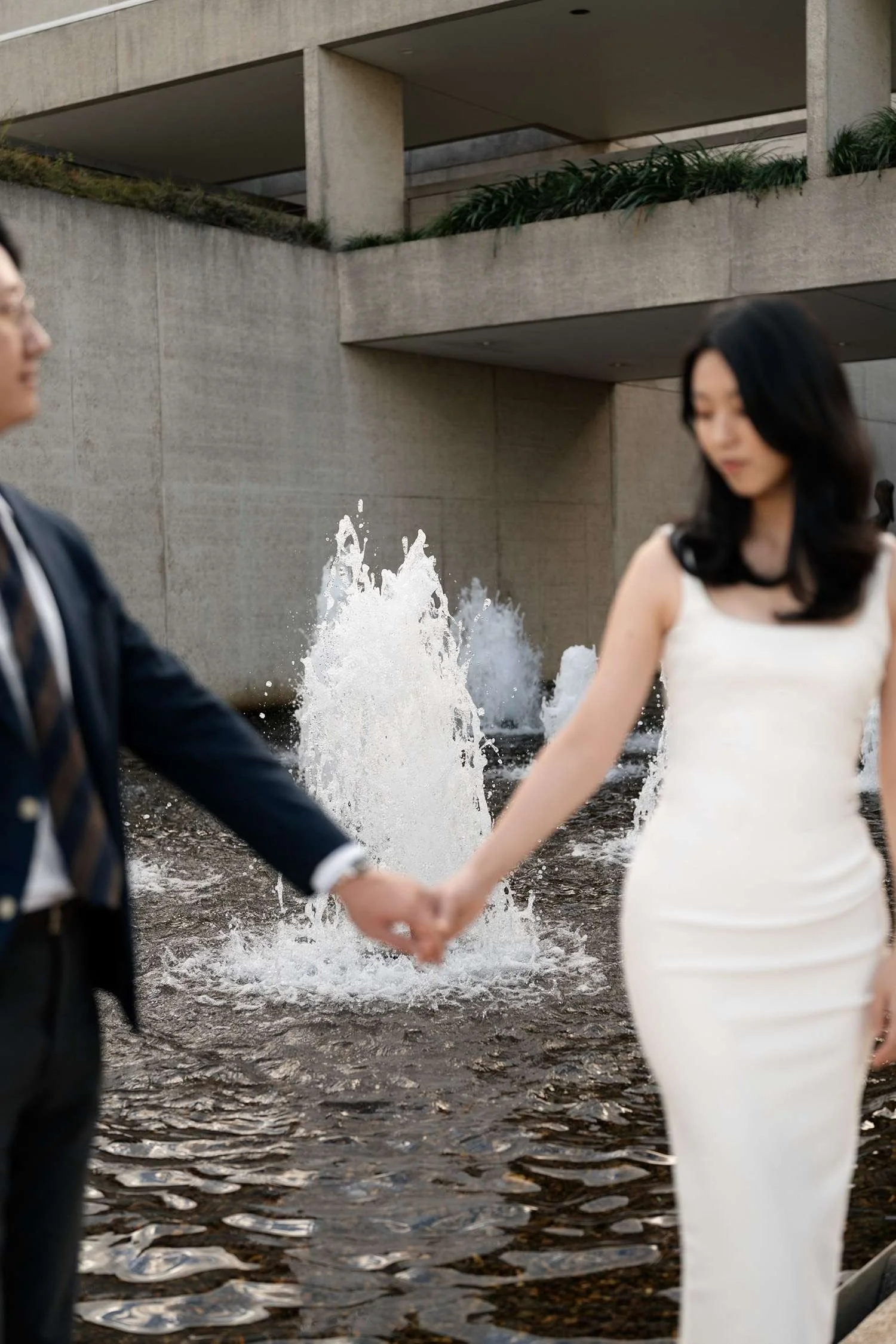Kath and Dave holding hands, standing together with a beautiful water fountain in the background, capturing a serene and romantic moment amidst the flowing water.