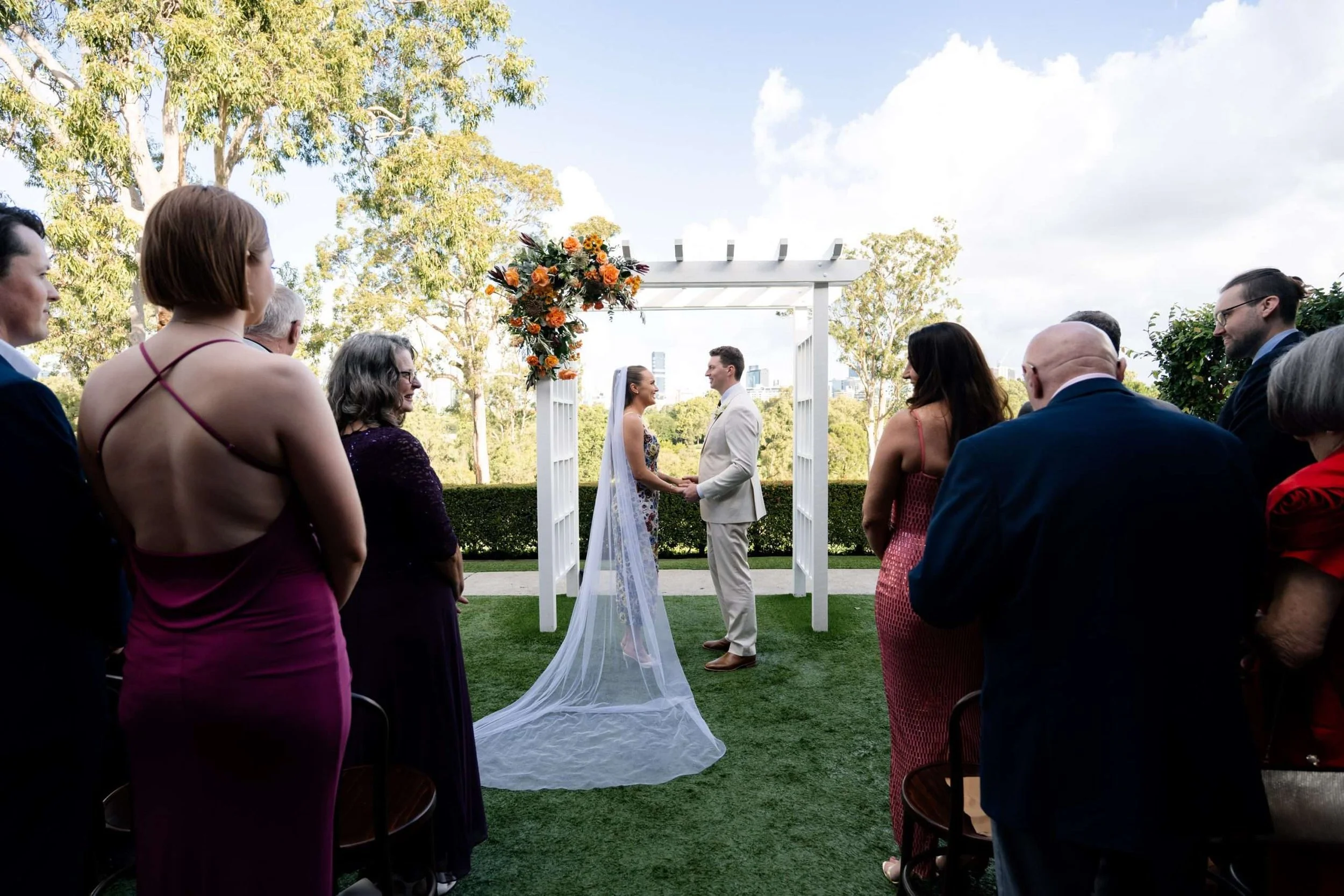 Bride and groom holding hands, looking at each other in front of the arbor, while their mothers smile at one another in Victoria park