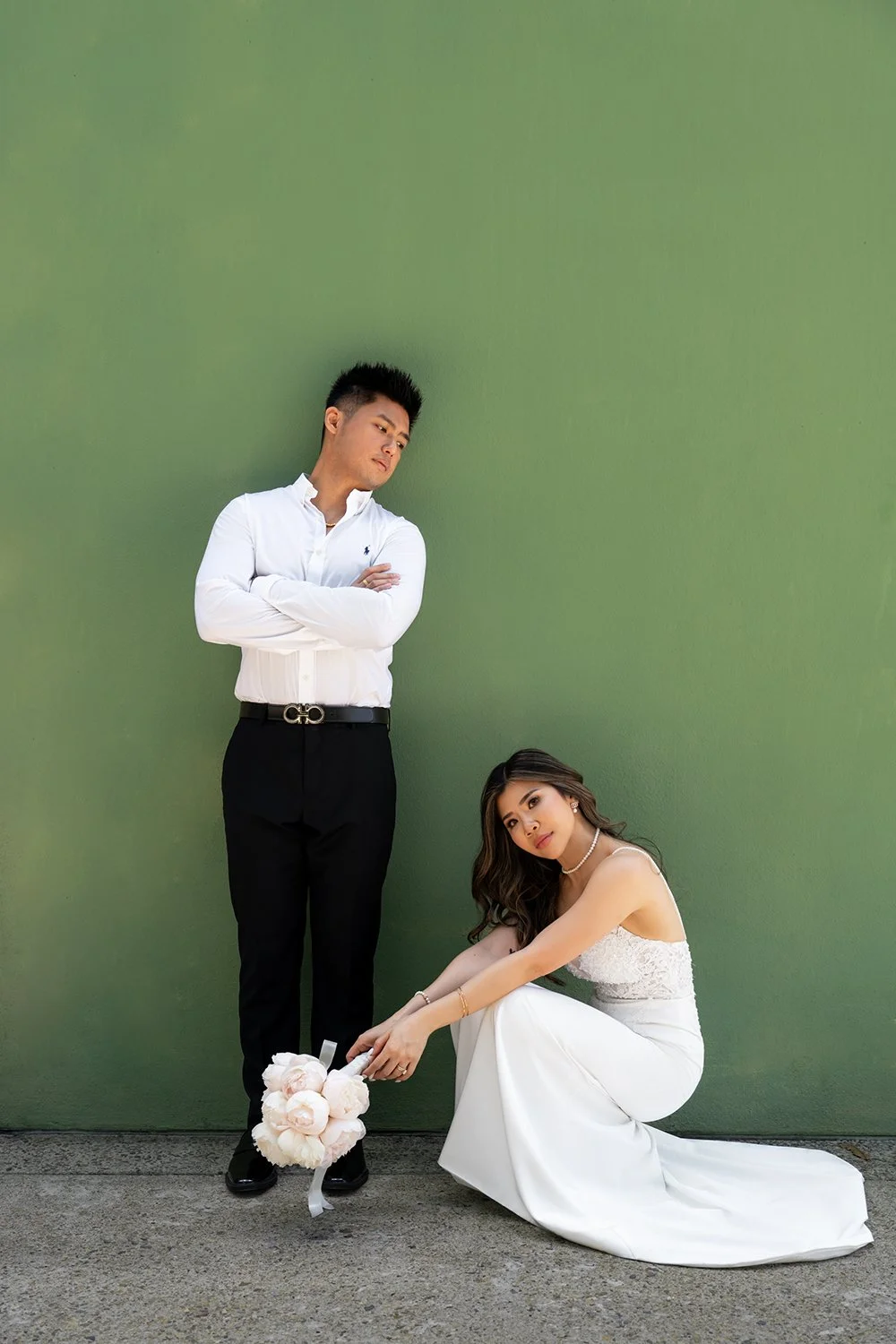 Bride Tiffy in a squat position, looking directly at the camera with a confident expression, while the groom Kai stands beside her, casually leaning against a green wall. The contrast in their poses adds a modern and stylish touch to this unique wedd
