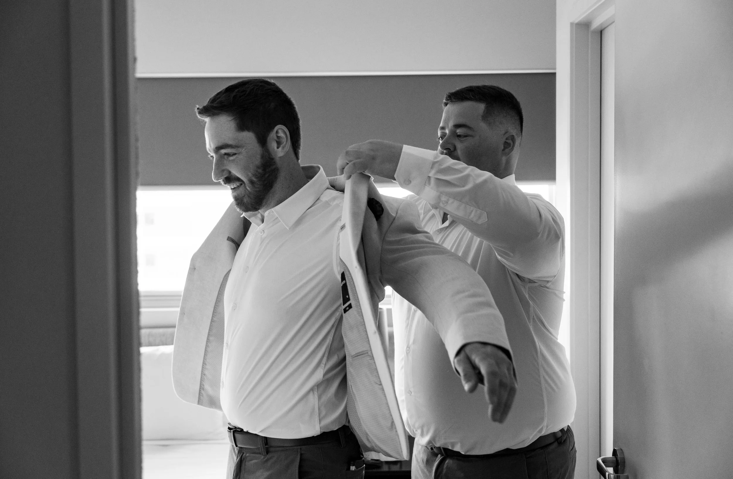 Best man helping groom Thomas put on his jacket, offering support and attention to detail as they prepare for the wedding ceremony