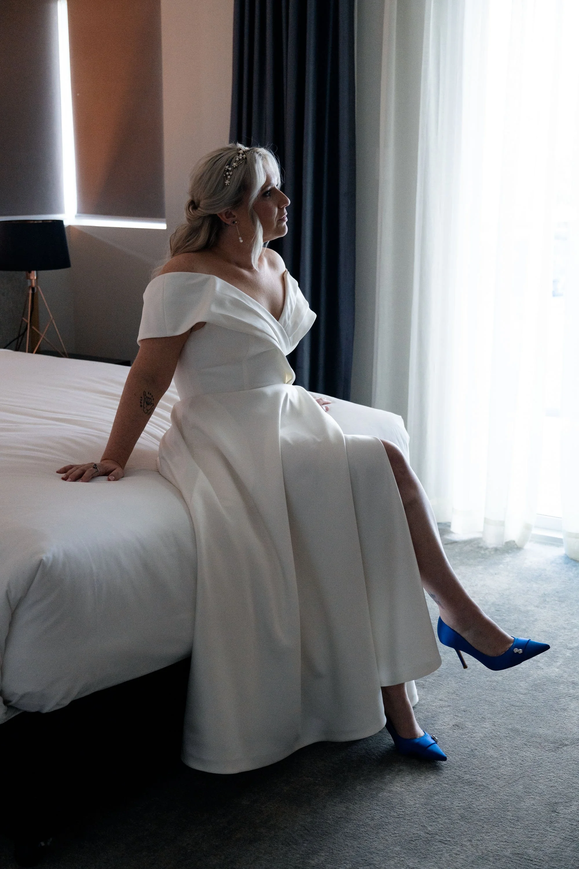 Bride Kyra resting her hands on the bed, looking up with a serene and contemplative expression. The soft lighting and gentle ambiance create a peaceful, intimate moment, capturing her beauty and the calm before the wedding