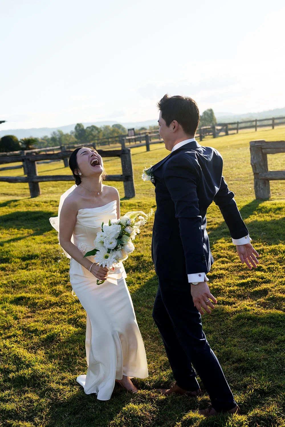 The bride Flora throws her head back in laughter while the groom Eric swings his arms behind him, sharing a carefree and joyful moment.