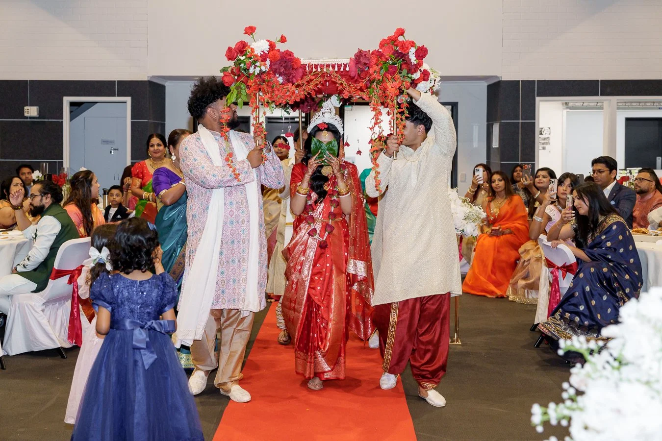 four males holding a floral Mandap and Amrita in the centre using 2 green leaves cover her eyes and they walking together