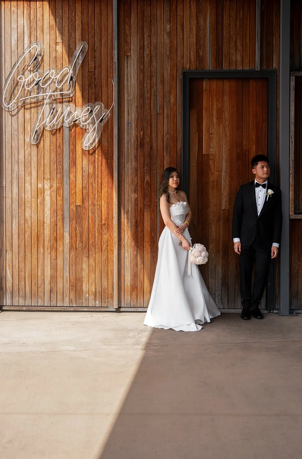 Bride Tiffy and groom Kai standing next to each other, with the bride looking at the Brisbane wedding photographer, while the groom leans casually in a relaxed pose. A rustic wooden wall serves as the backdrop, creating a natural and intimate vibe in