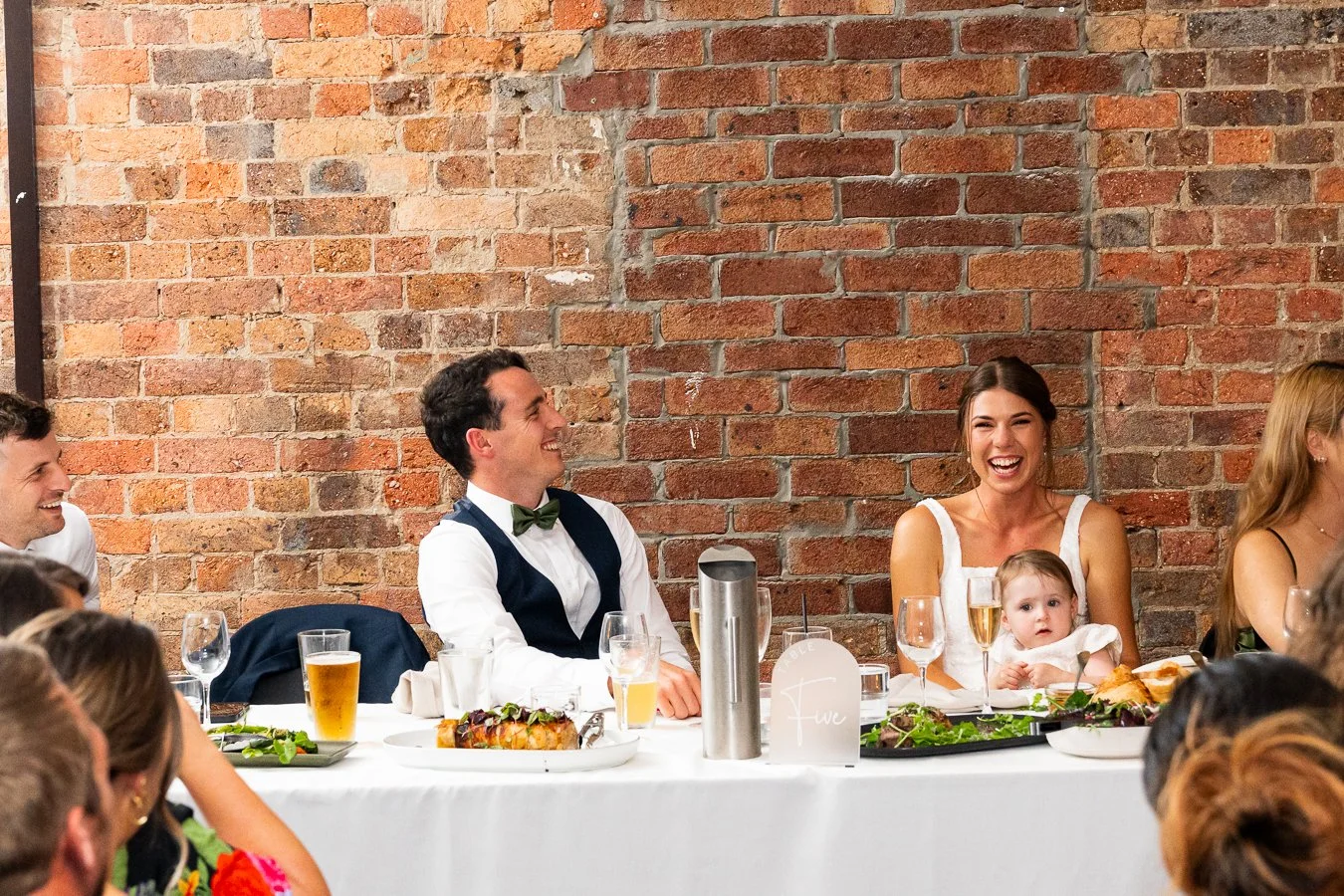 Giordan and Sonya laughing while sitting behind the table with a baby on Sonya's leg