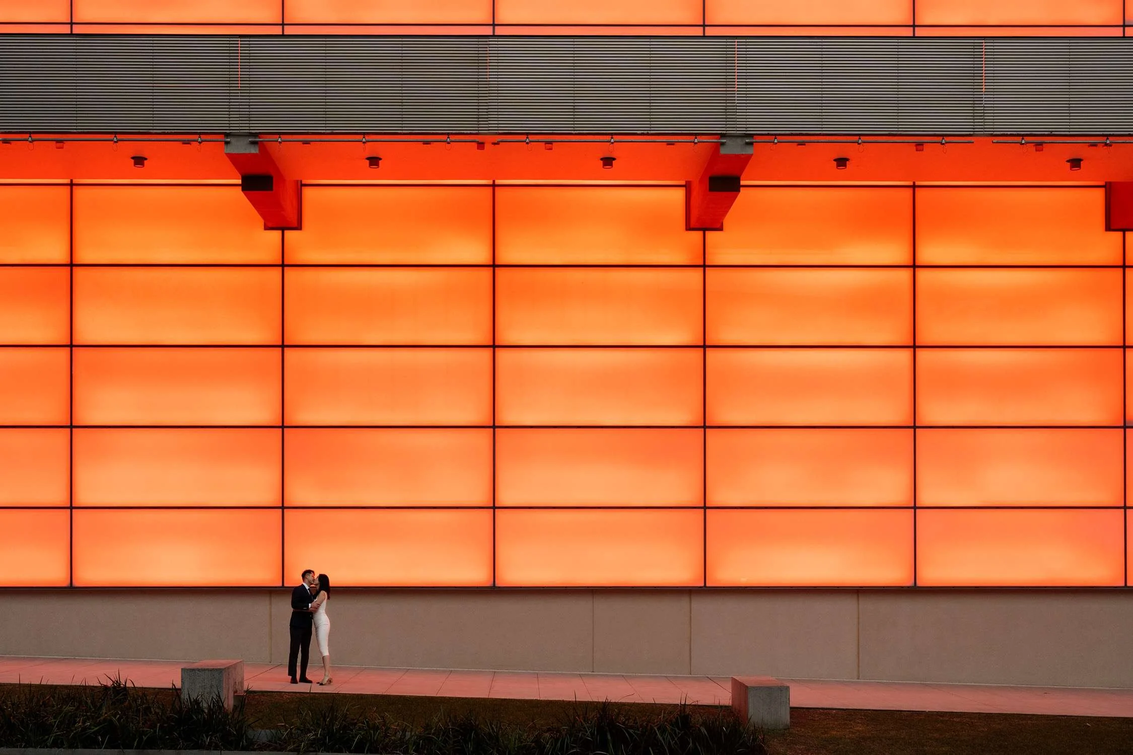 Kath and Dave sharing a kiss outside the Queensland Art Museum, illuminated by the vibrant glow of an orange LED wall behind them, creating a modern and romantic scene