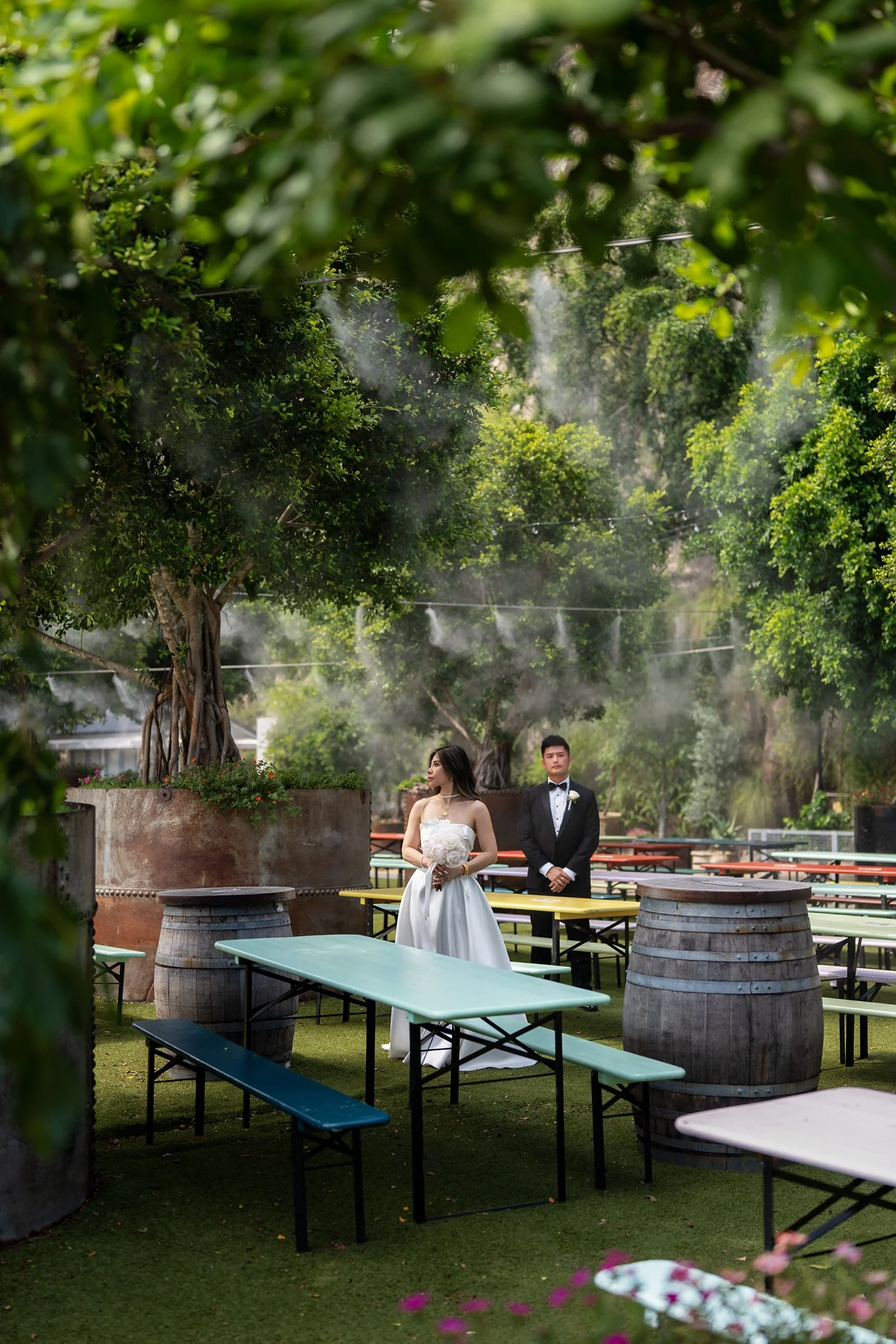 Bride Tiffy and groom Kai standing in an outdoor restaurant, surrounded by lush trees with a refreshing water spray in the air on a hot summer day. Colorful tables are scattered around them, adding a vibrant and joyful atmosphere to the romantic sett