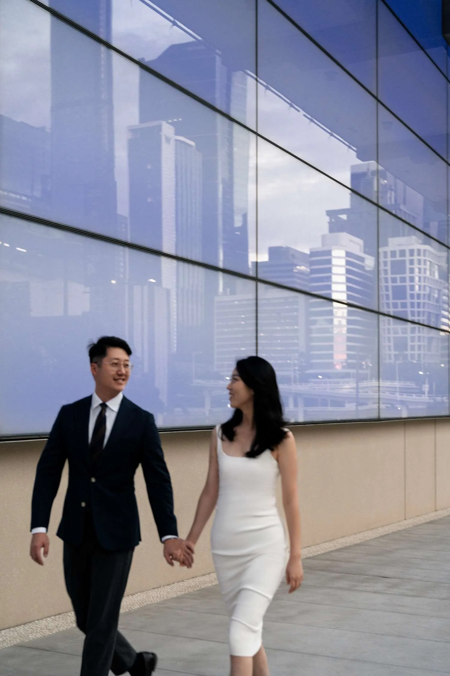 Kath and Dave holding hands as they walk together, gazing lovingly at each other outside the Queensland Art Museum, surrounded by its architectural charm