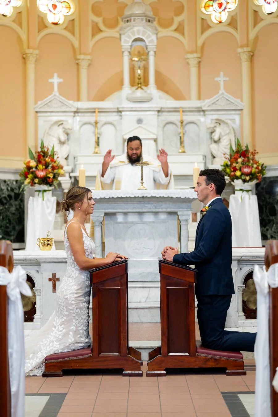 The bride and groom kneel before a small table, facing each other, while the priest stands at the altar in the distance.