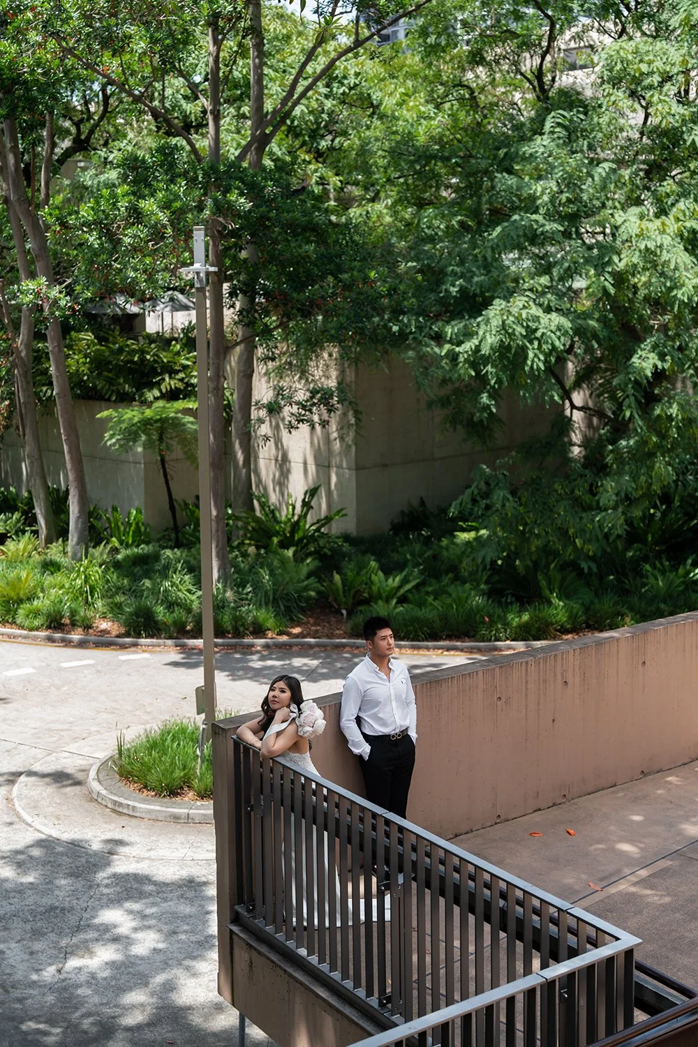 Bride Tiffy standing next to a fence, gazing directly at the Brisbane wedding photographer, while the groom Kai stands a step away, looking off into the distance. The bride wears a beautiful wedding gown, and the groom is dressed in a classic suit, c