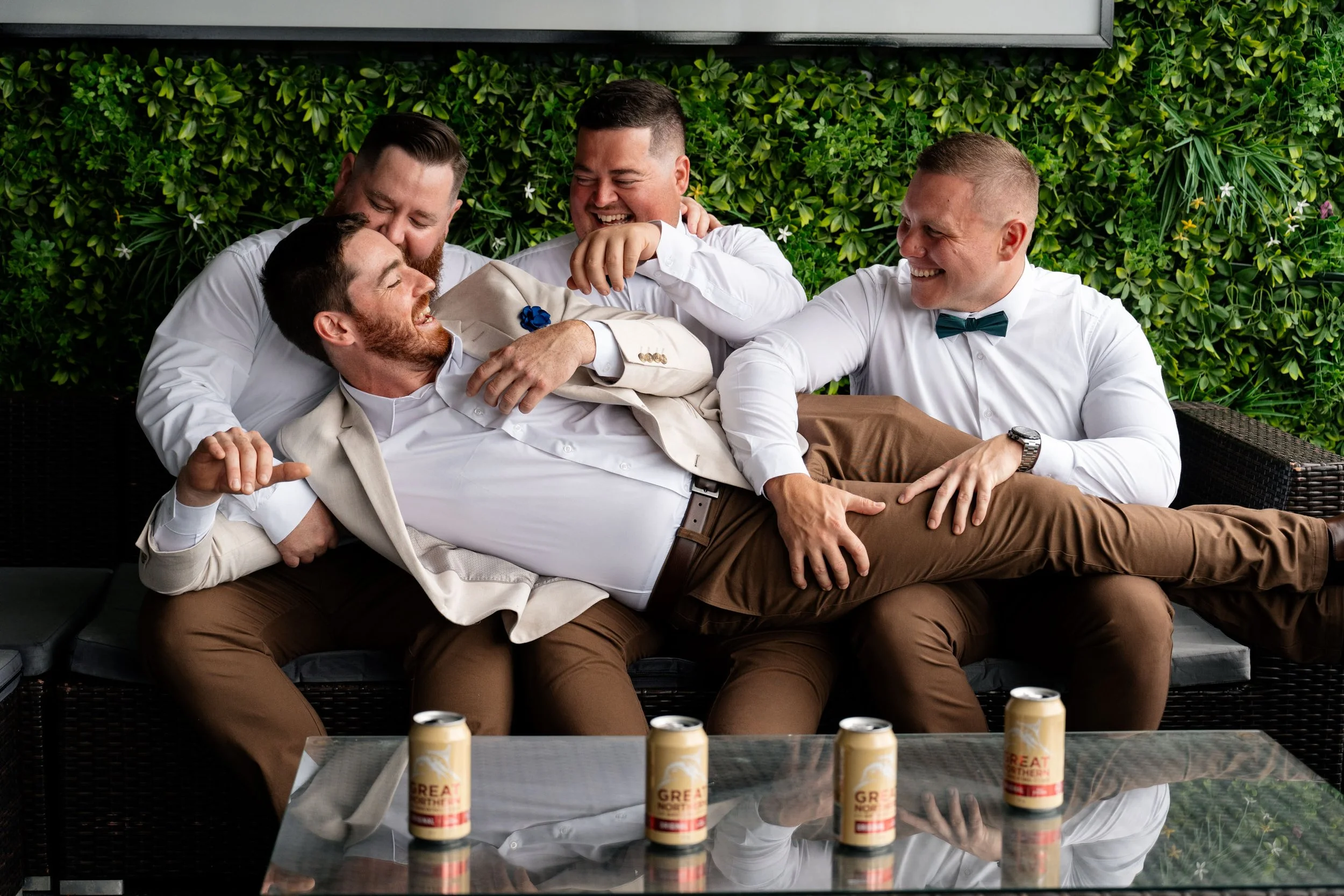 Groom Thomas lying down on his groomsmen's leg, sharing a fun and lighthearted moment as they all laugh together during the wedding celebration