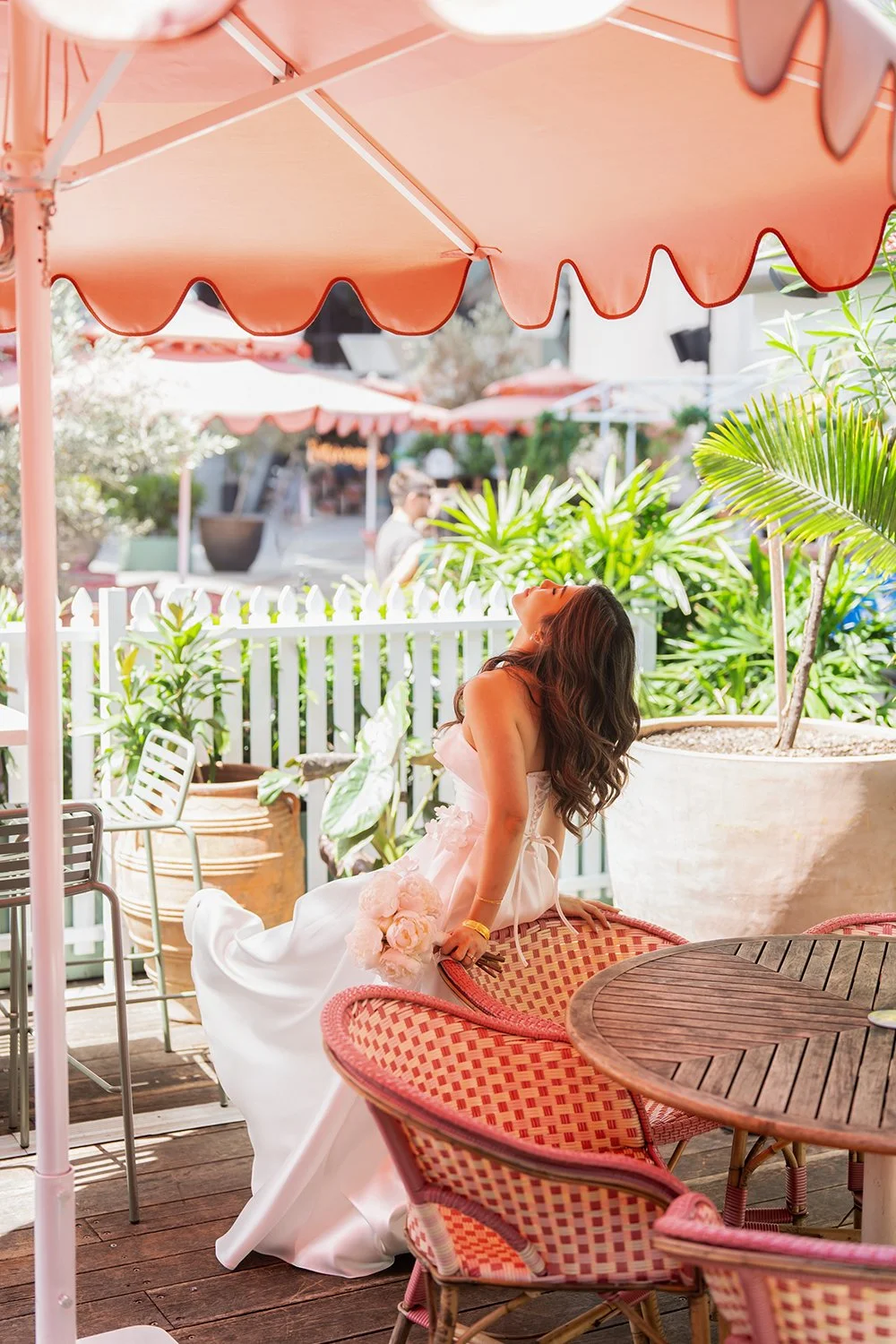 Bride Tiffy sitting backward on a chair with her arms resting on the backrest, her head tilted upward in a relaxed pose. She is under a large pink umbrella, wearing her white wedding gown, with soft natural light creating a playful and modern bridal 